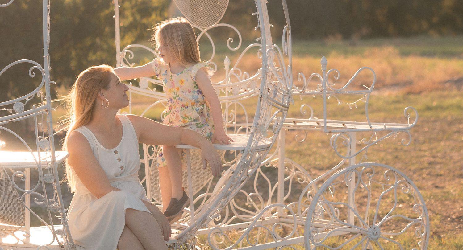 Mother and daughter sitting in a white ornate carriage outdoors in golden sunlight.