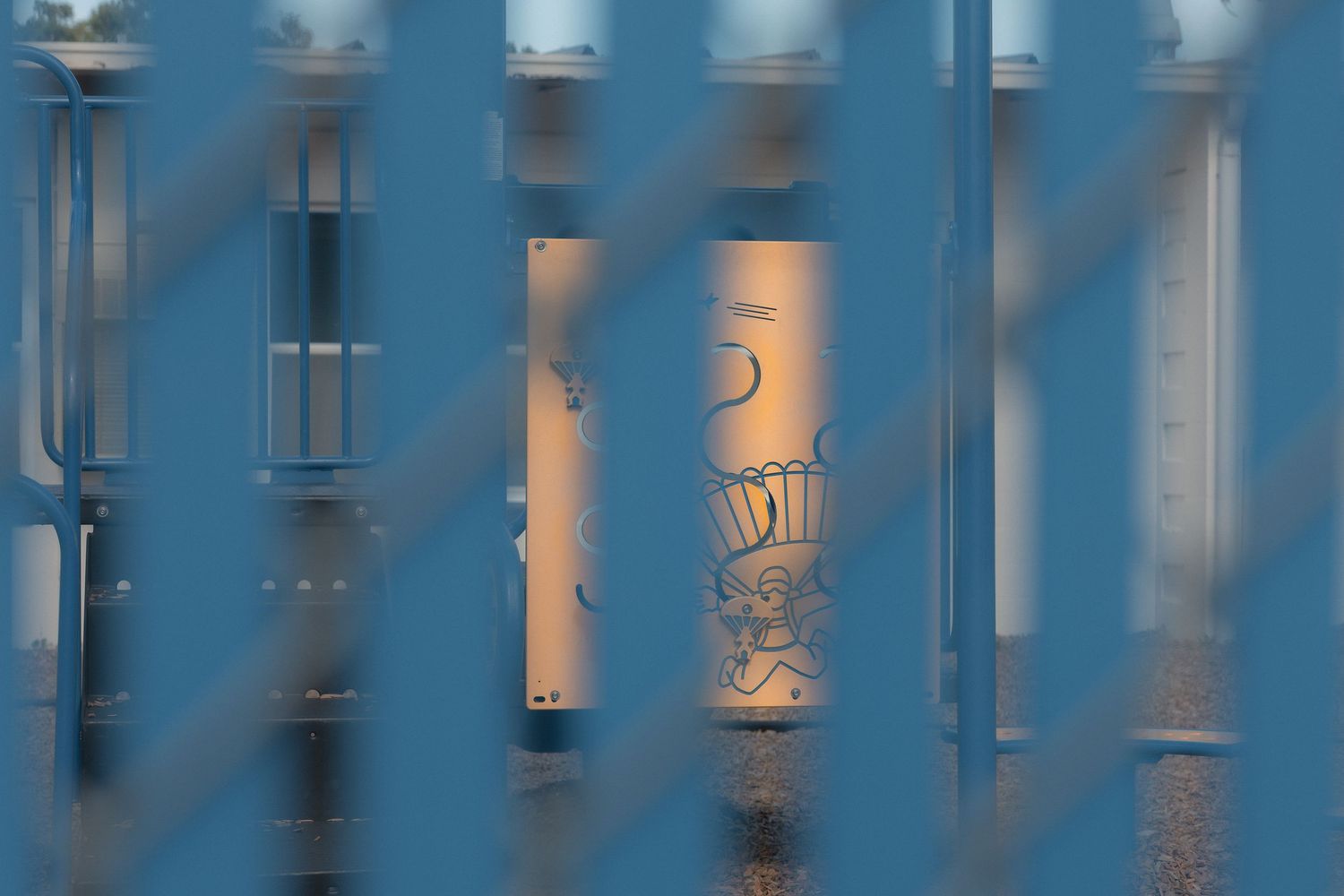Graffiti art visible through blue metal fence slats on a wall, photographed at dusk with warm golden light.