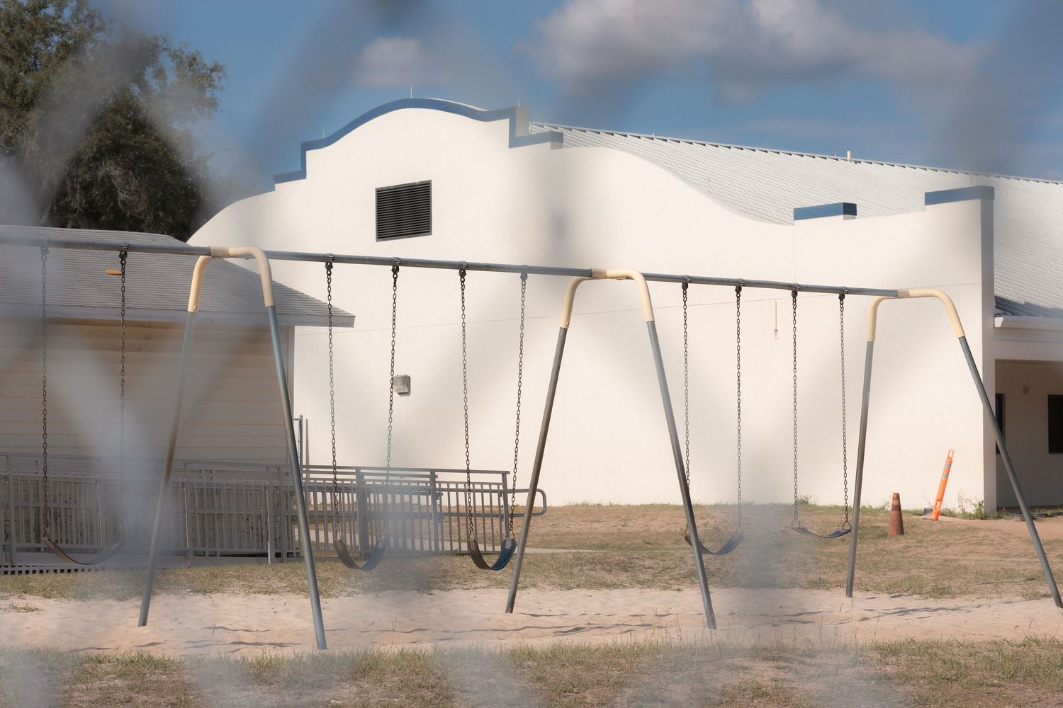 Empty metal swing set on sandy ground in front of a white school building on a sunny day.