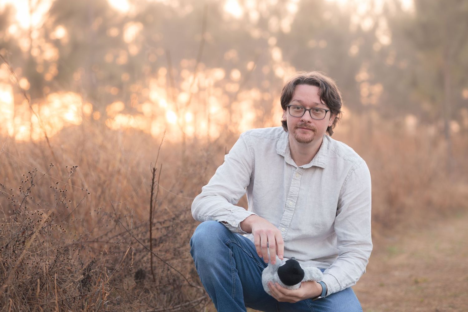A photographer in a white sweater and jeans sits in a golden field during sunset with professional camera equipment.