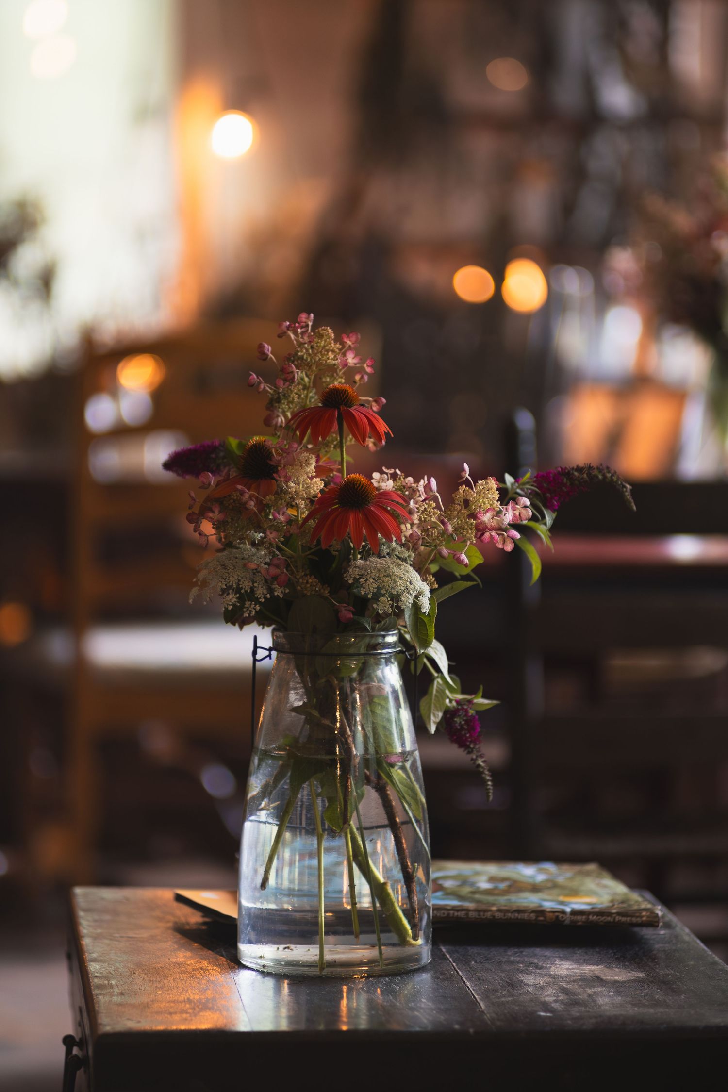 A rustic glass jar vase with wildflowers on a dark wooden table in a warmly lit restaurant setting.