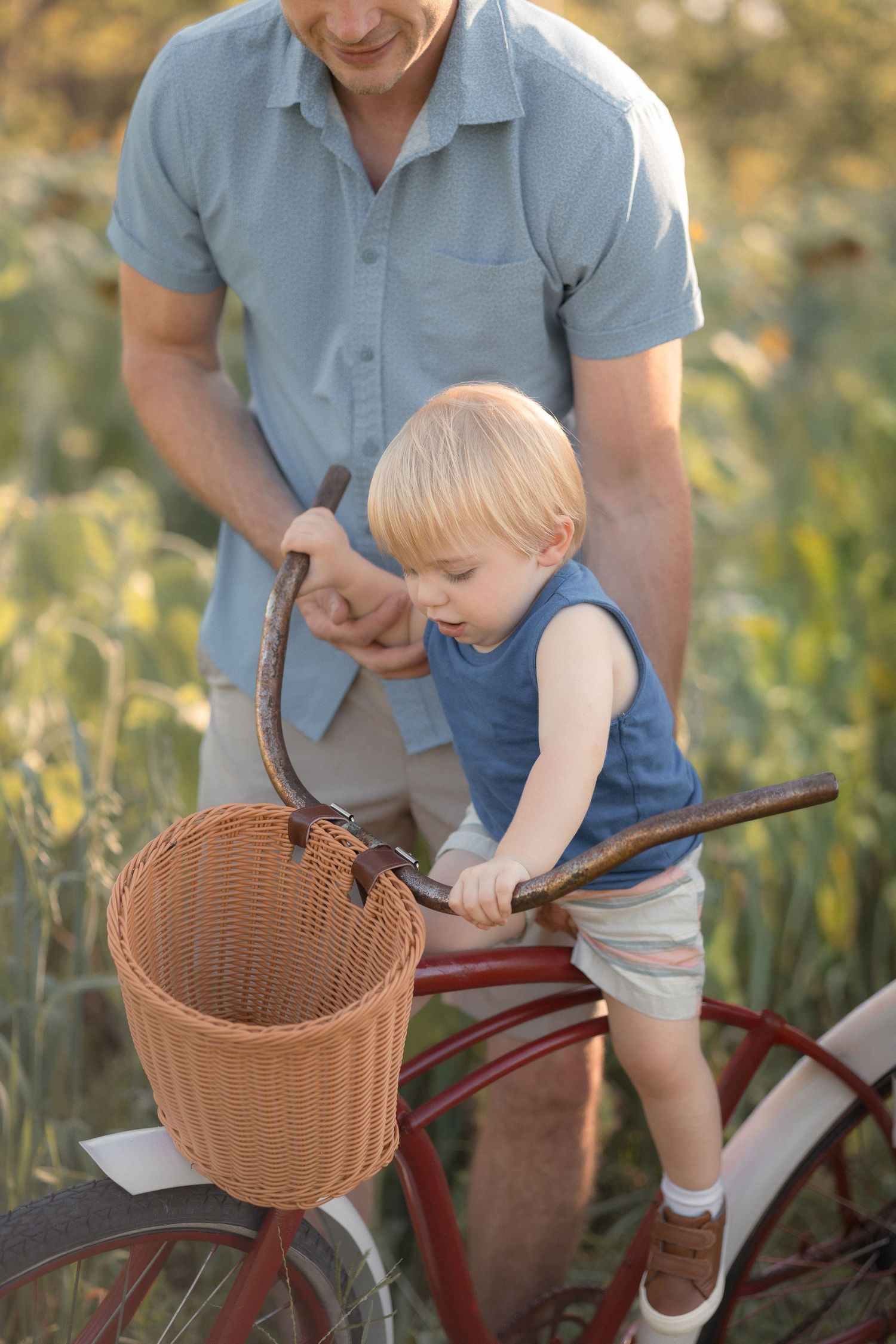 Father helping toddler boy hold bicycle handlebars with wicker basket in sunny green field.