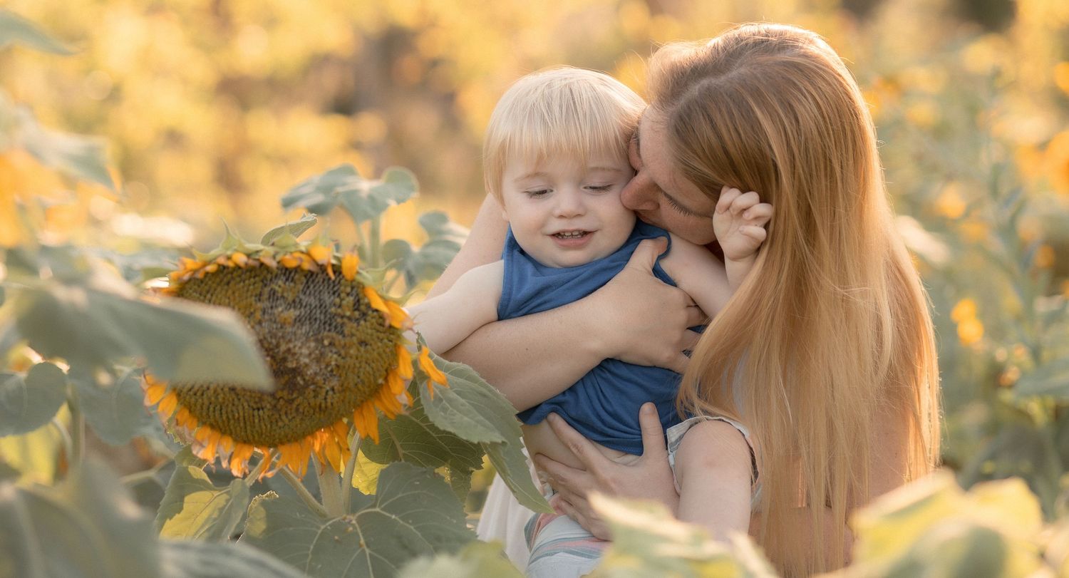 Mother kissing her toddler son in a sunny sunflower field, golden light illuminating their joyful outdoor moment.
