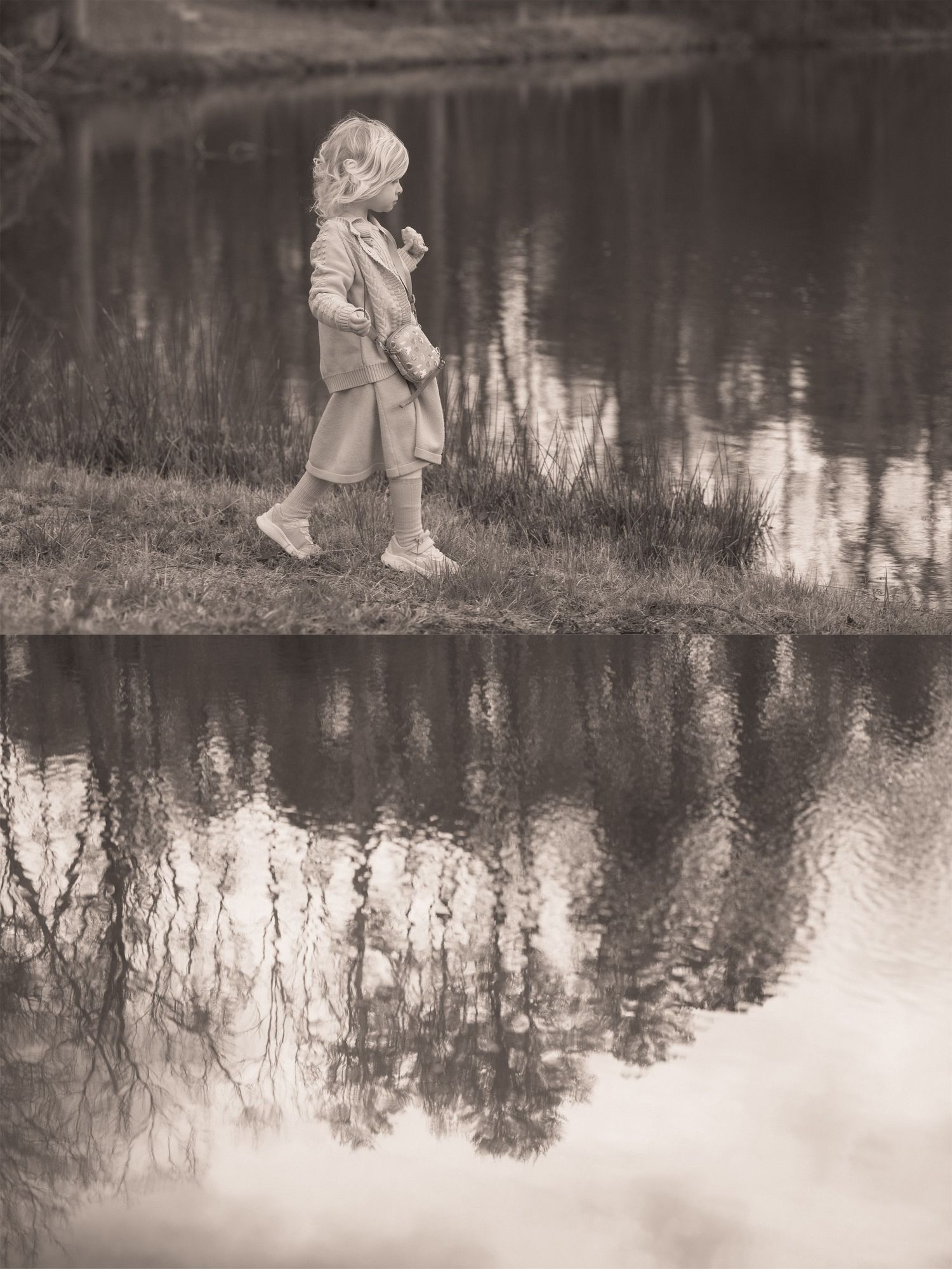 Sepia-toned photo of a young child walking along a reflective pond edge, with tree reflections mirrored in the still water.