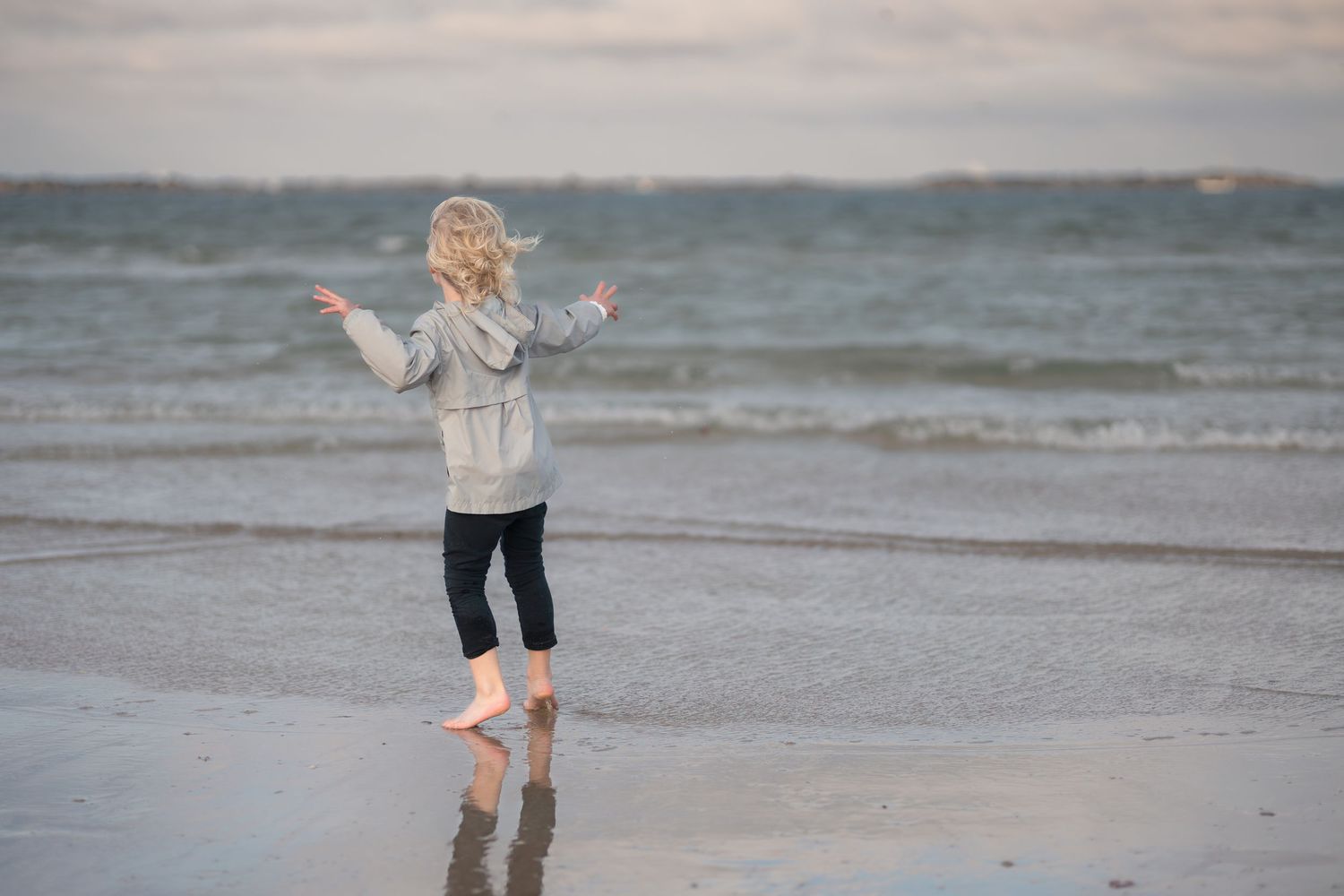 A person in a cream sweater walks along a misty beach shoreline at sunset.