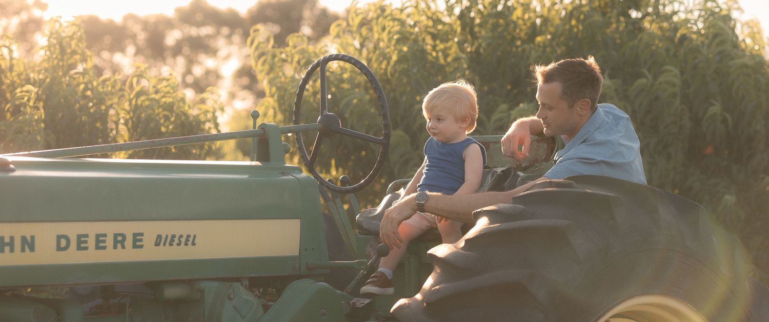 Father and baby sitting together on a green John Deere tractor in a sunlit farm field.