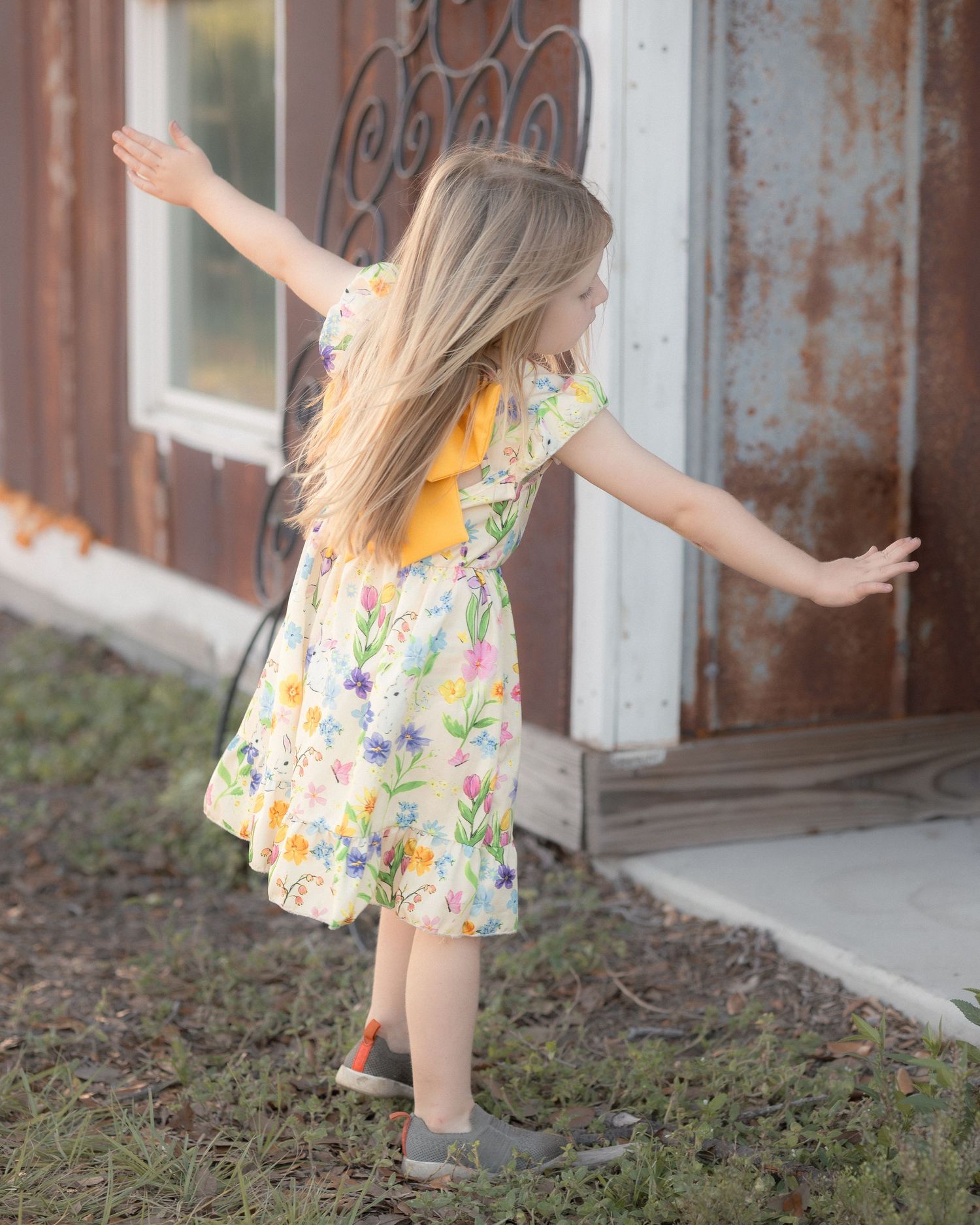 Young girl in floral dress with yellow bodice twirling outdoors near rustic building.