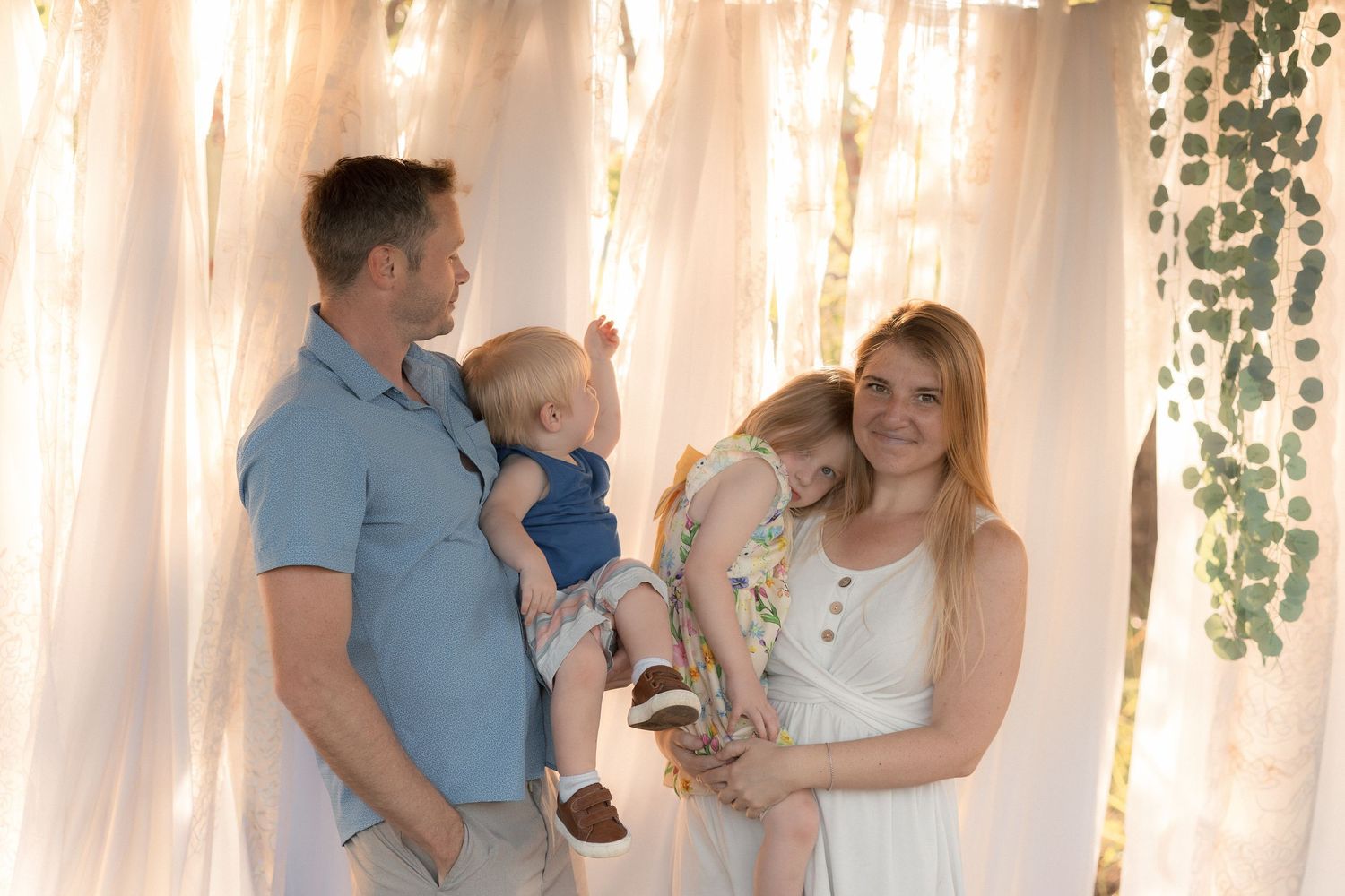Family of four posing outdoors, parents holding young children, smiling warmly in soft natural light near draped white curtains.