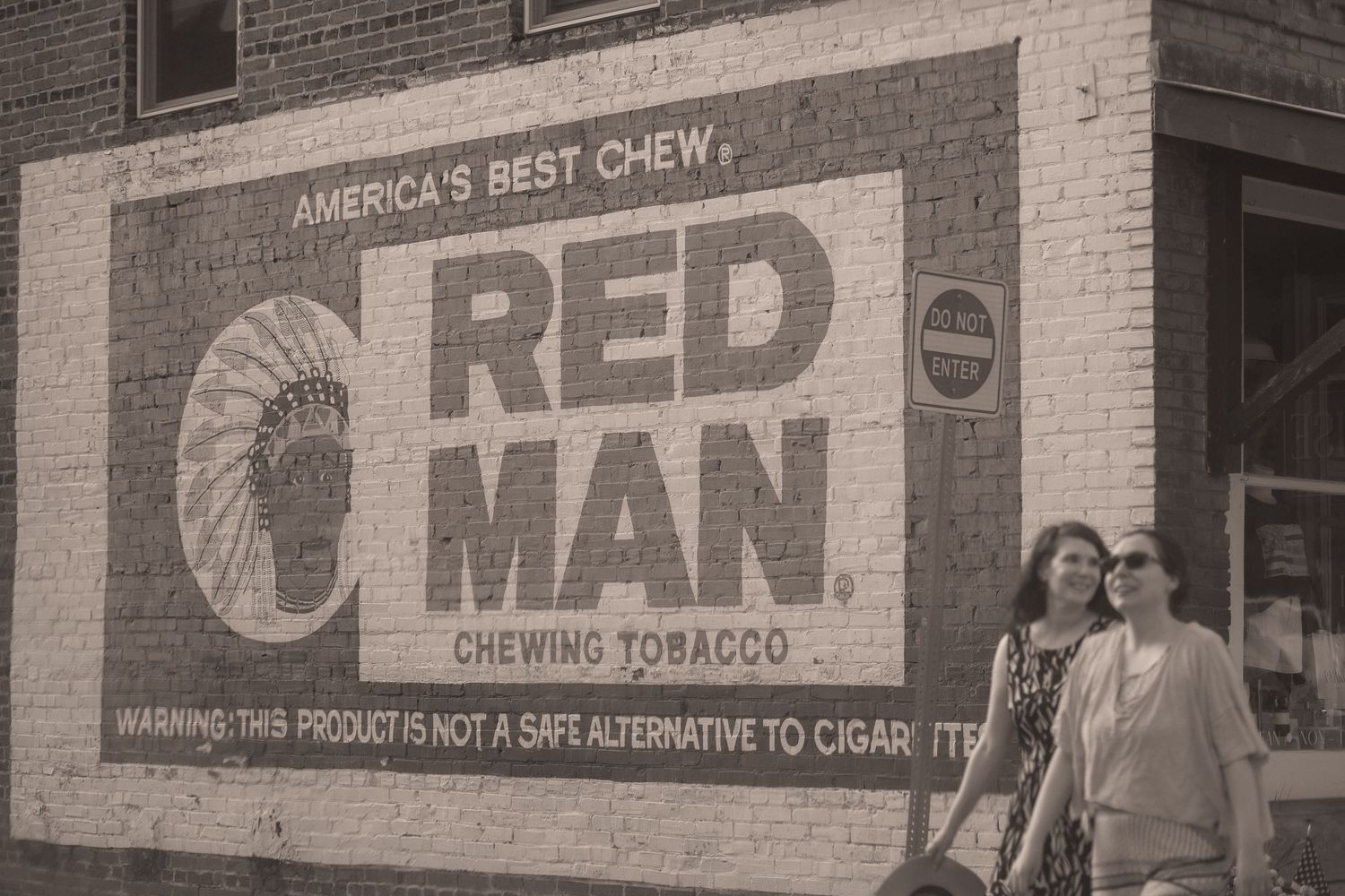 Two women stand in front of a vintage Red Man Chewing Tobacco painted brick wall advertisement in a small town.
