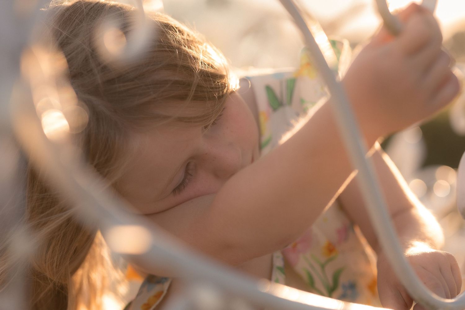 Young girl with blonde hair playing outdoors in golden sunlight, wearing a floral dress among white metal railings.