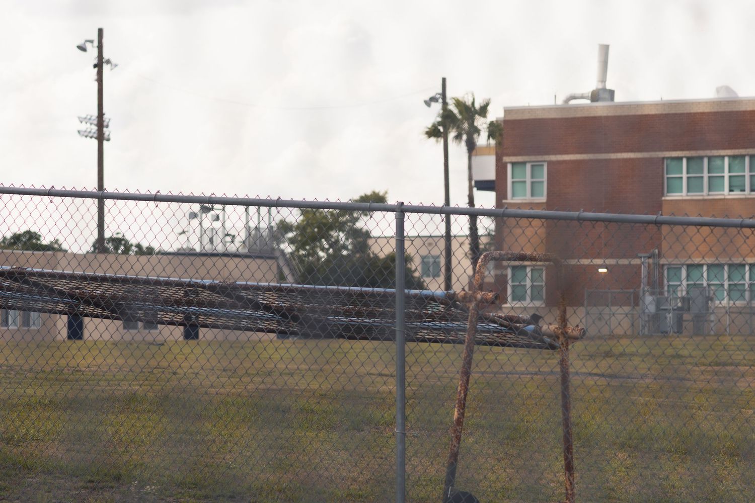 Chain-link fence surrounds a brick school building with palm trees in background on an overcast day.