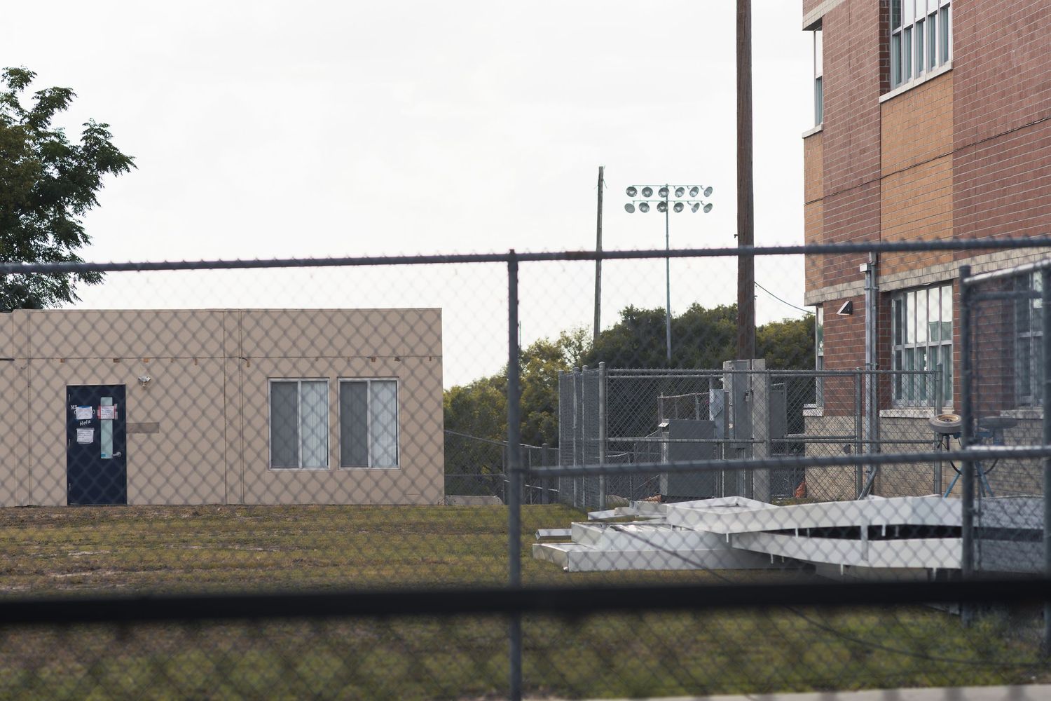 Chain-link fence surrounds an industrial building complex with signage visible in the background on a cloudy day.