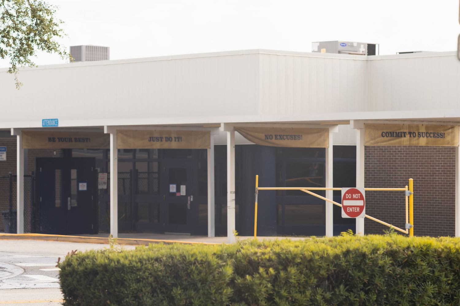 Exterior view of a commercial building with multiple dark glass doors, yellow safety barriers, and a do not enter sign.