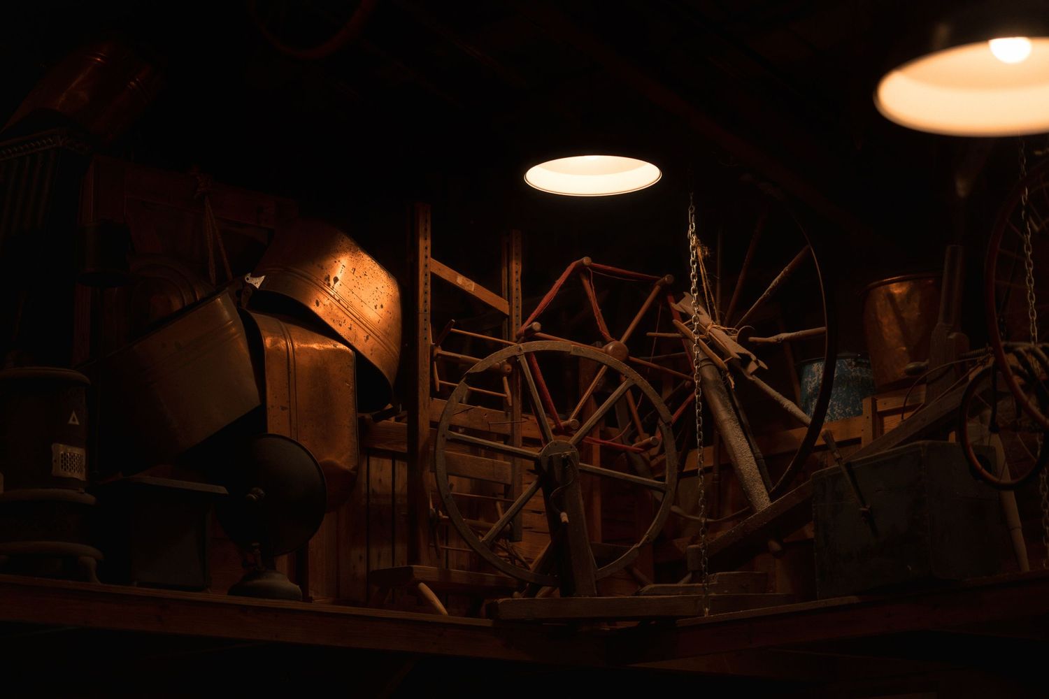 Dimly lit barn interior with vintage wooden wagon wheel and antique farm equipment under warm pendant lights.