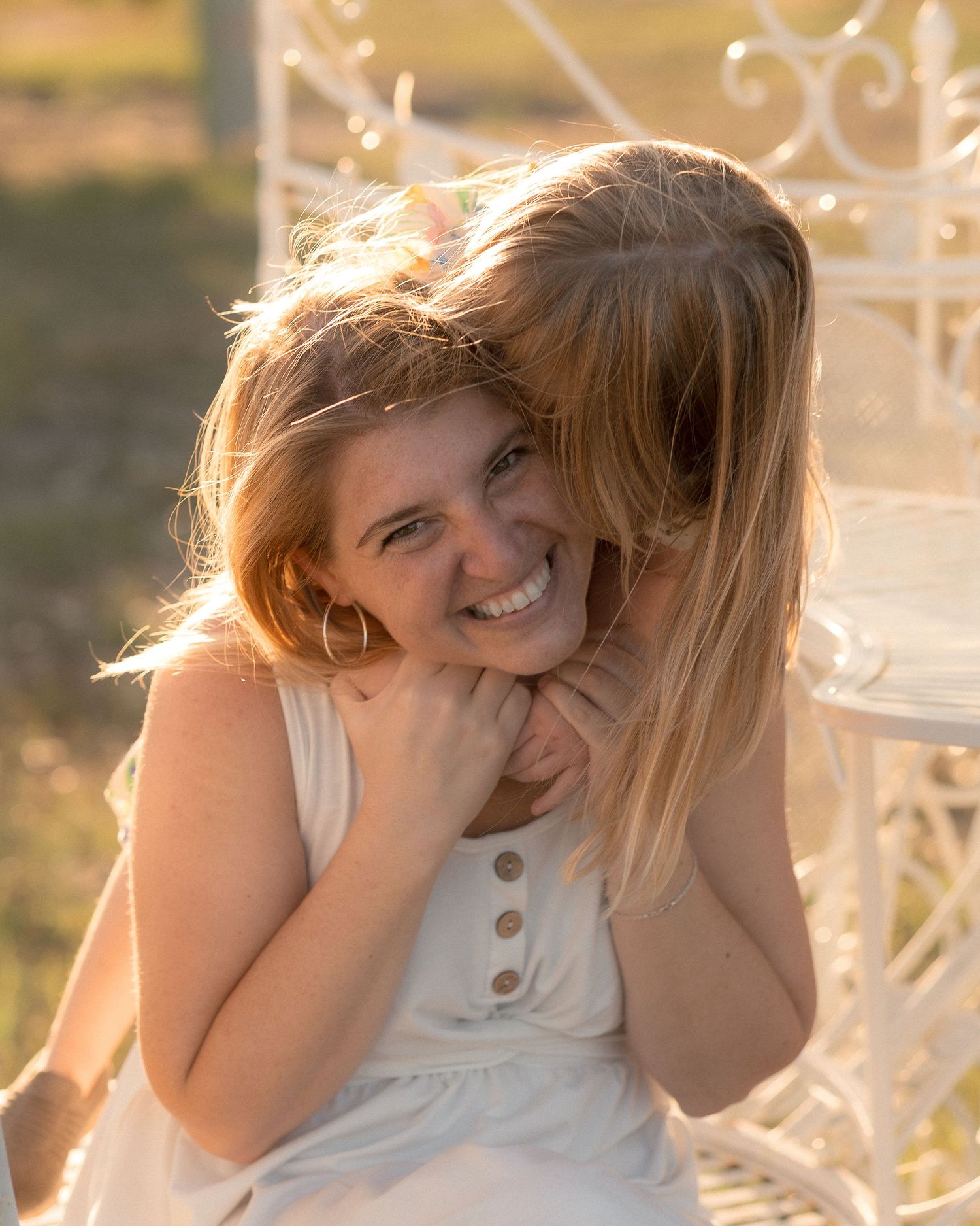 A smiling woman hugs a young blonde girl on a white bench outdoors in warm golden light.