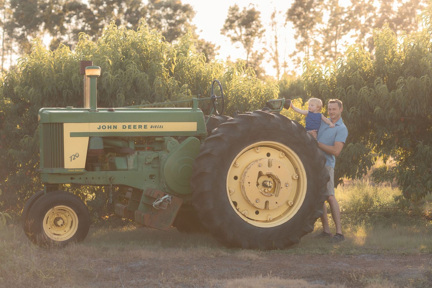 Farmer holding a child beside a vintage green John Deere tractor in a sunlit orchard at golden hour.