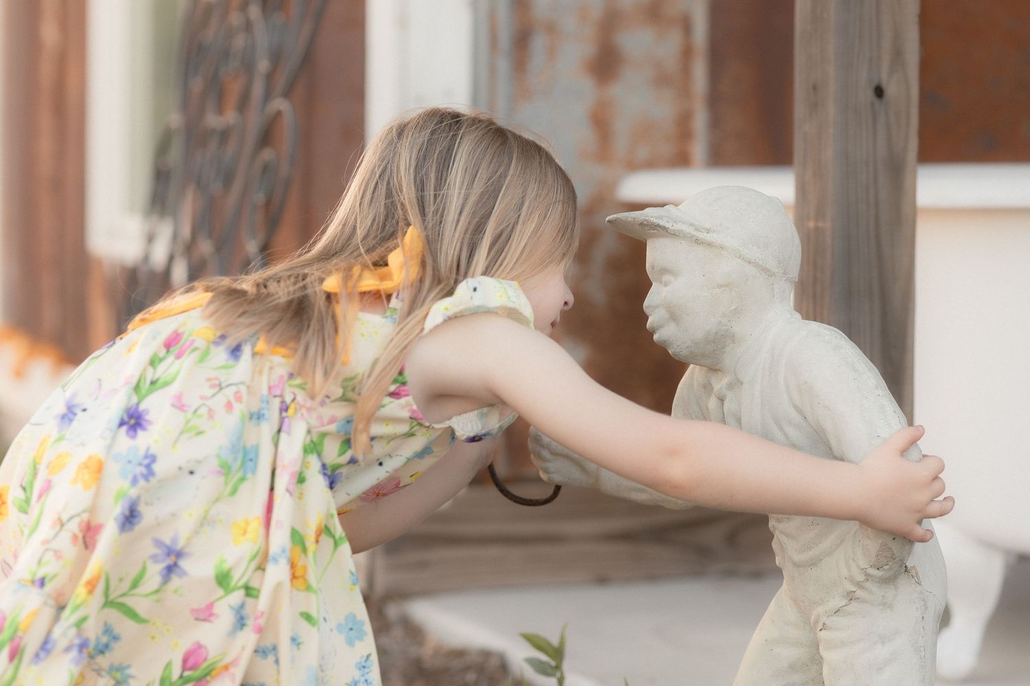 Young girl in floral dress leaning in to kiss a white stone garden statue of a child outdoors.
