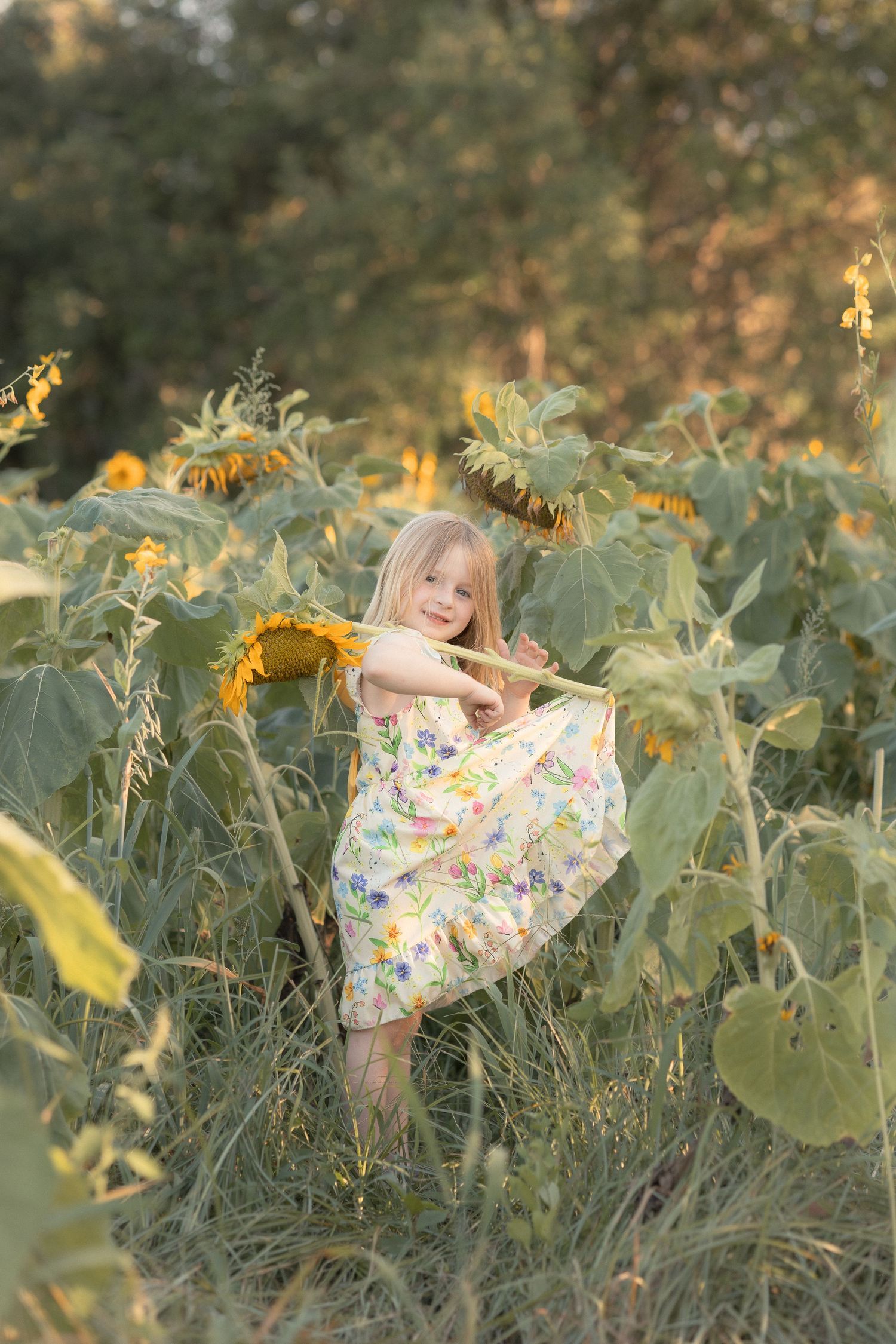 Young blonde girl in floral dress twirling joyfully among tall sunflowers in a golden field.