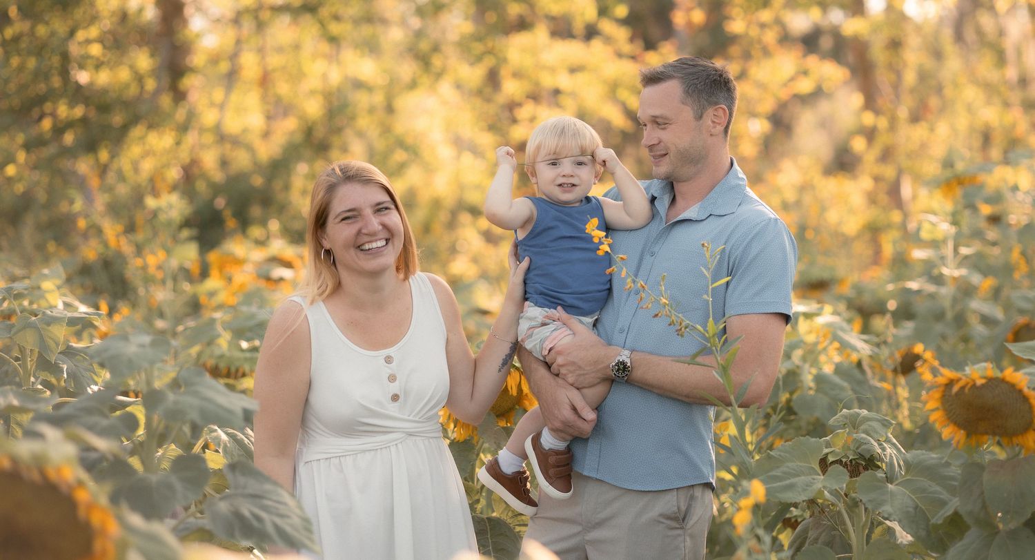 Happy family with toddler posing in sunflower field during golden hour outdoor portrait session.