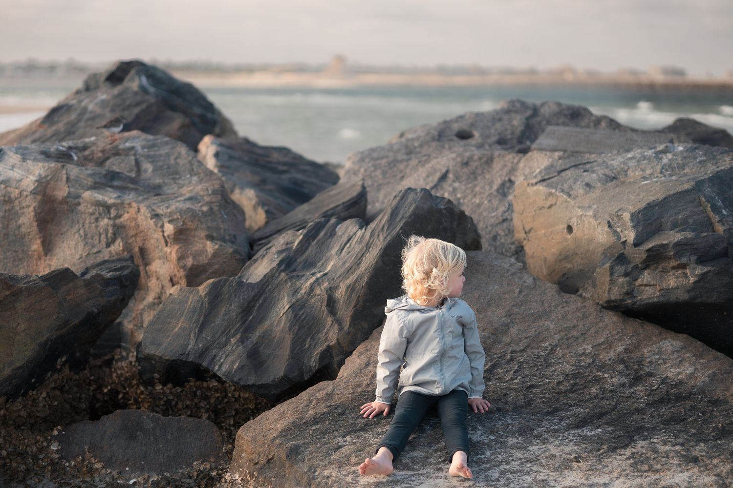 Small child in a gray sweater sits among large coastal rocks at sunset by the ocean.