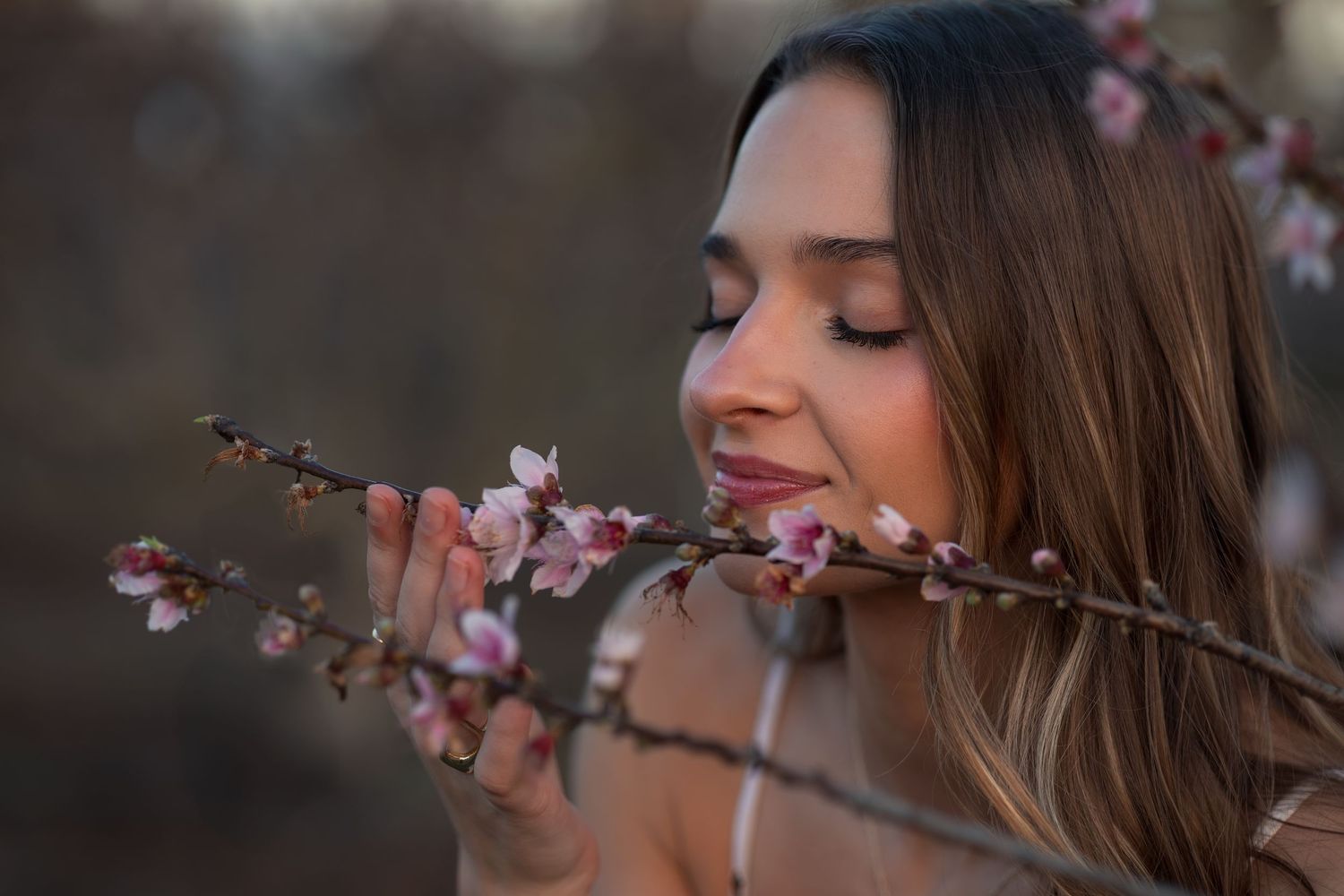 Woman with long brown hair smells pink cherry blossoms on a tree branch in spring.