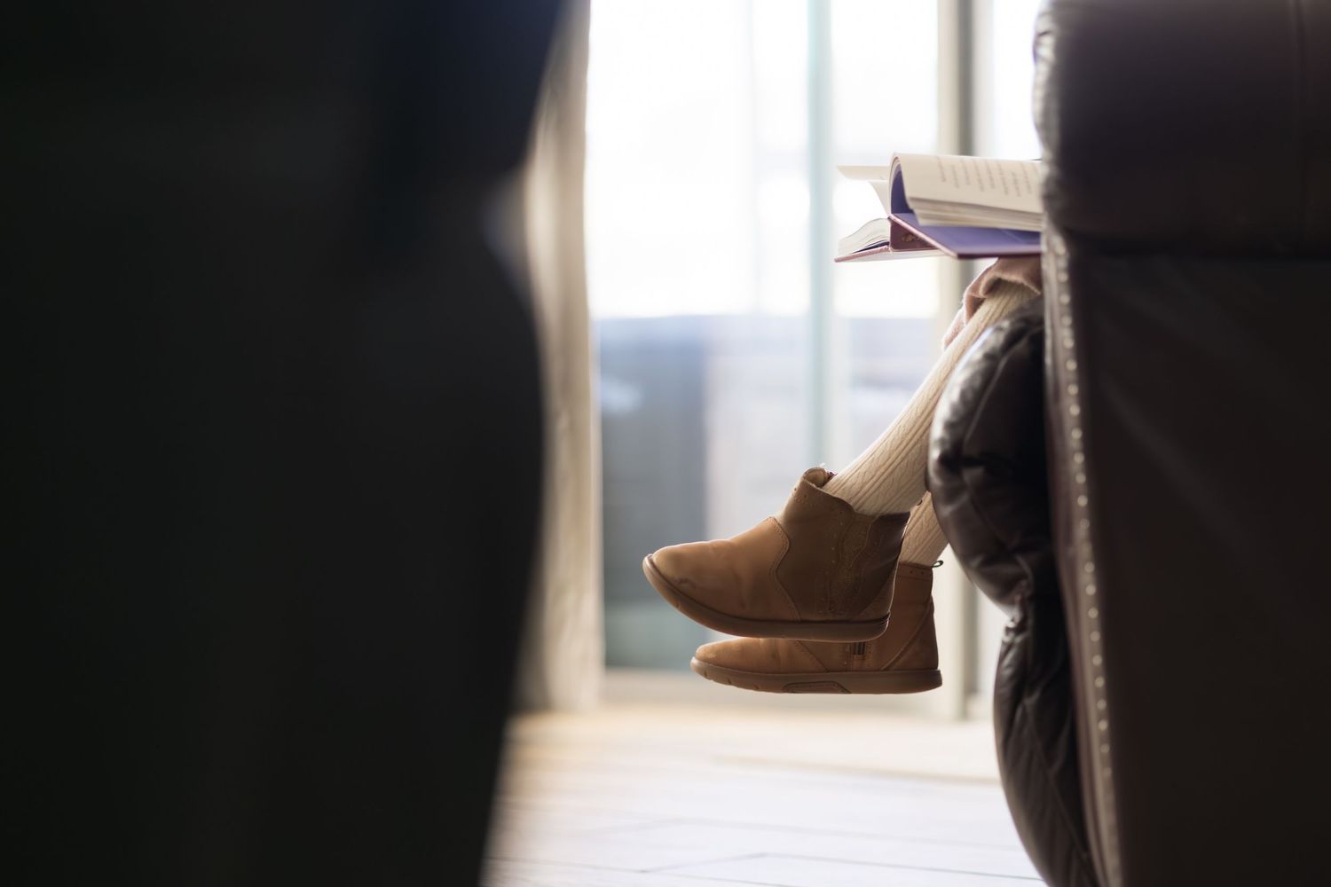 A brown leather boot sits near a window, casting a soft shadow on the wooden surface in natural light.