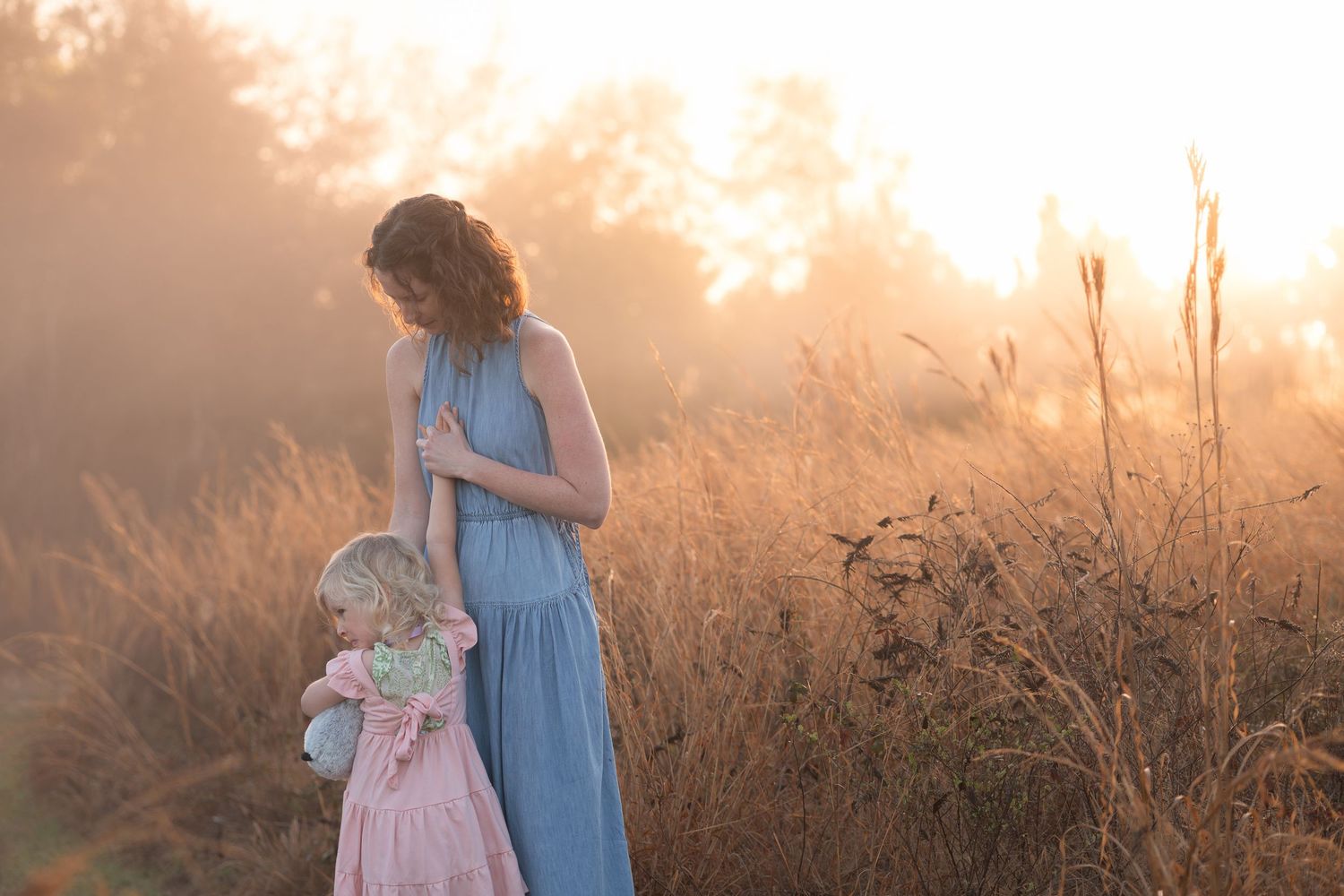 Two figures in soft blue and pink dresses stand in a golden sunlit meadow during sunset.