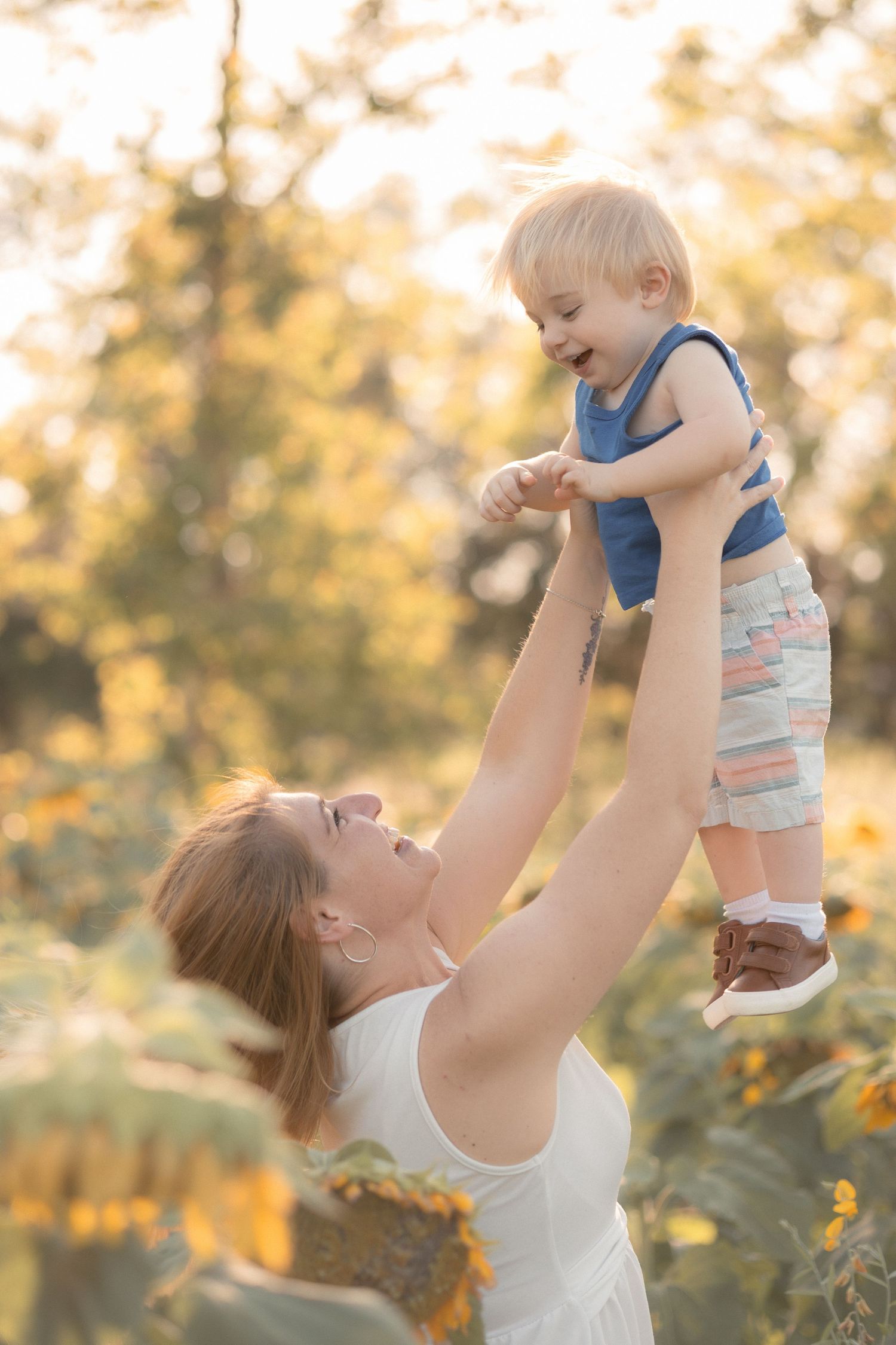 Mother lifting smiling toddler in the air outdoors in a sunflower field during golden hour.