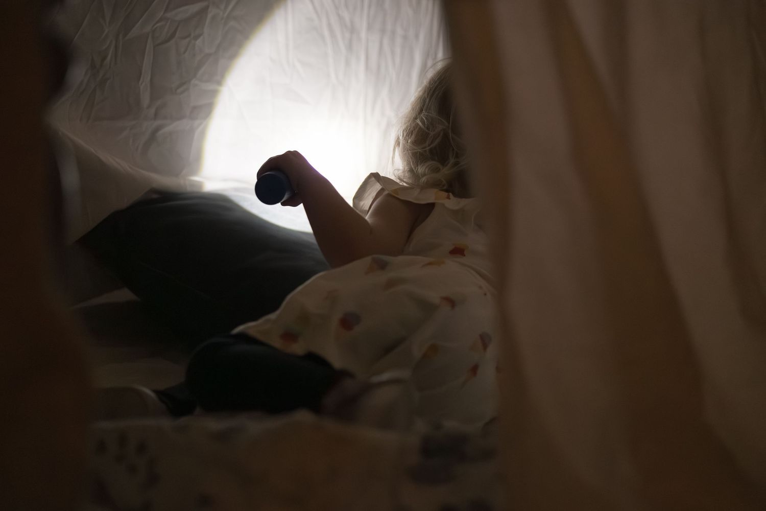 A black cat stretches and yawns while lounging on a bed with white sheets in dim lighting.