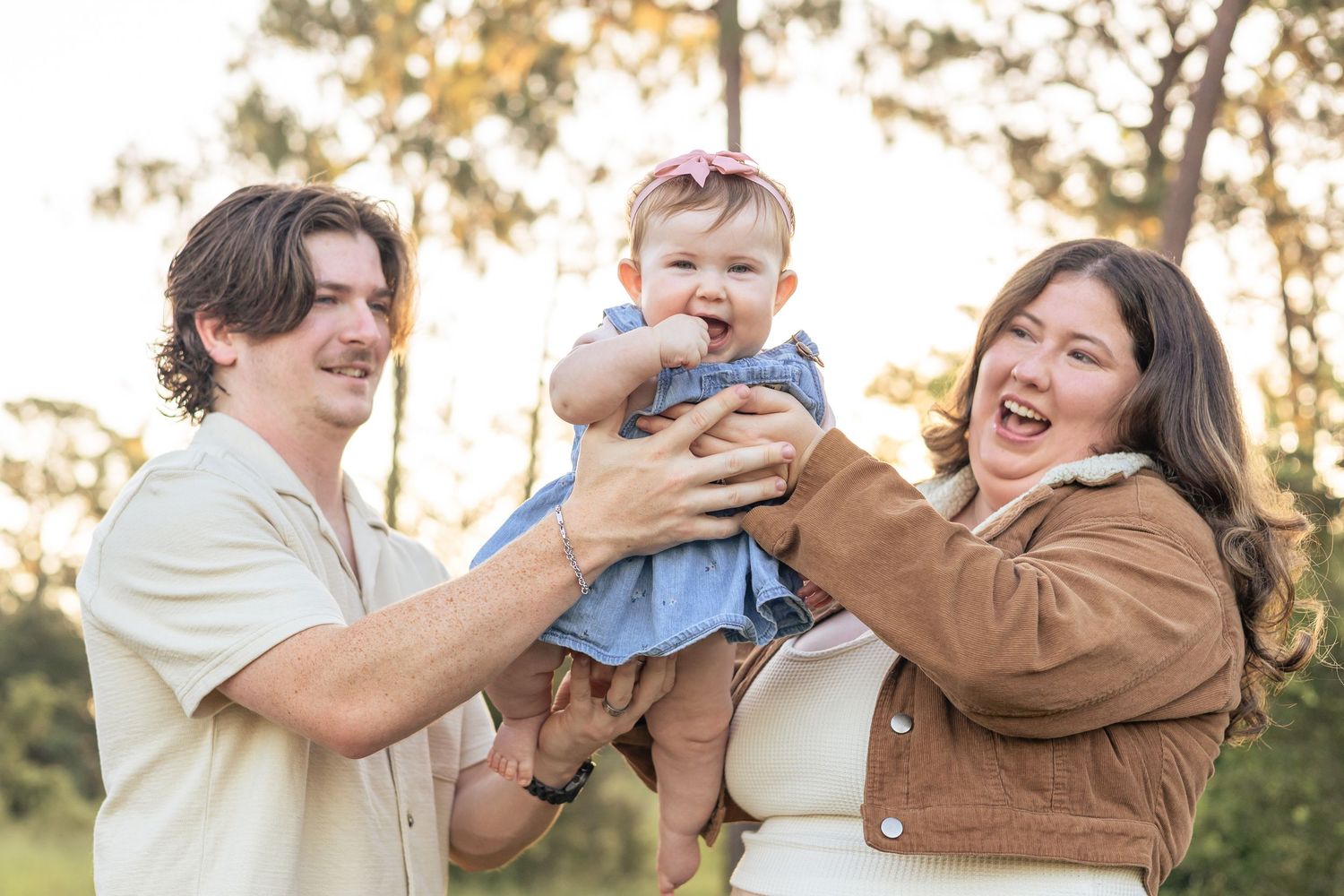 A family laughs and plays together during a golden hour outdoor photo session in a park setting.