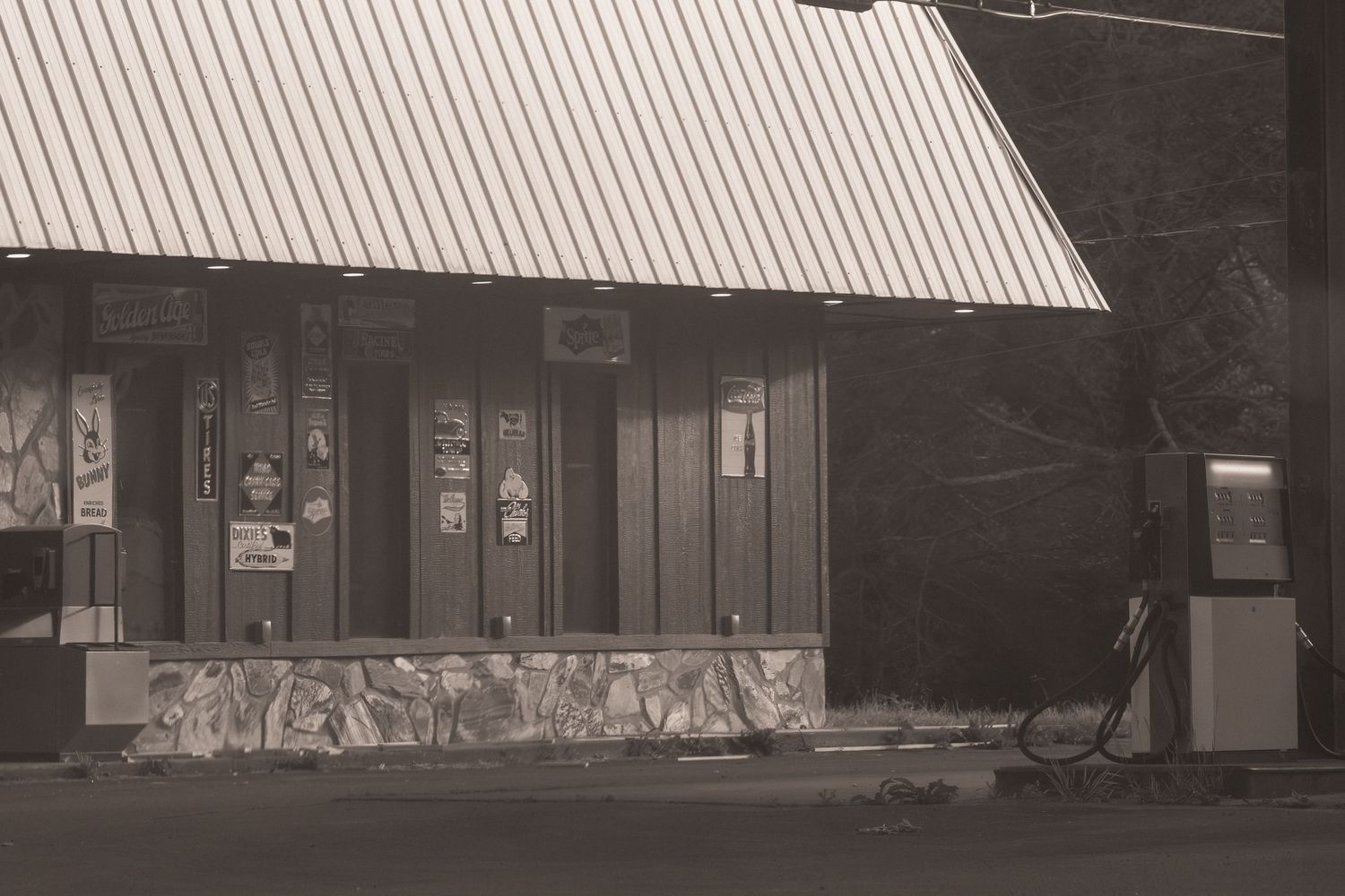 Sepia-toned photo of a vintage roadside building with metal roof, stone facade, and old gas pump at dusk.