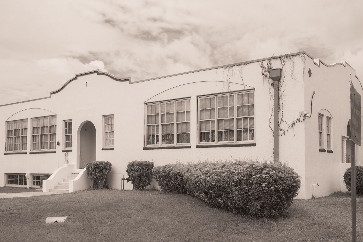 Black and white photo of a vintage white stucco school building with arched entrance, large windows, and trimmed hedges.