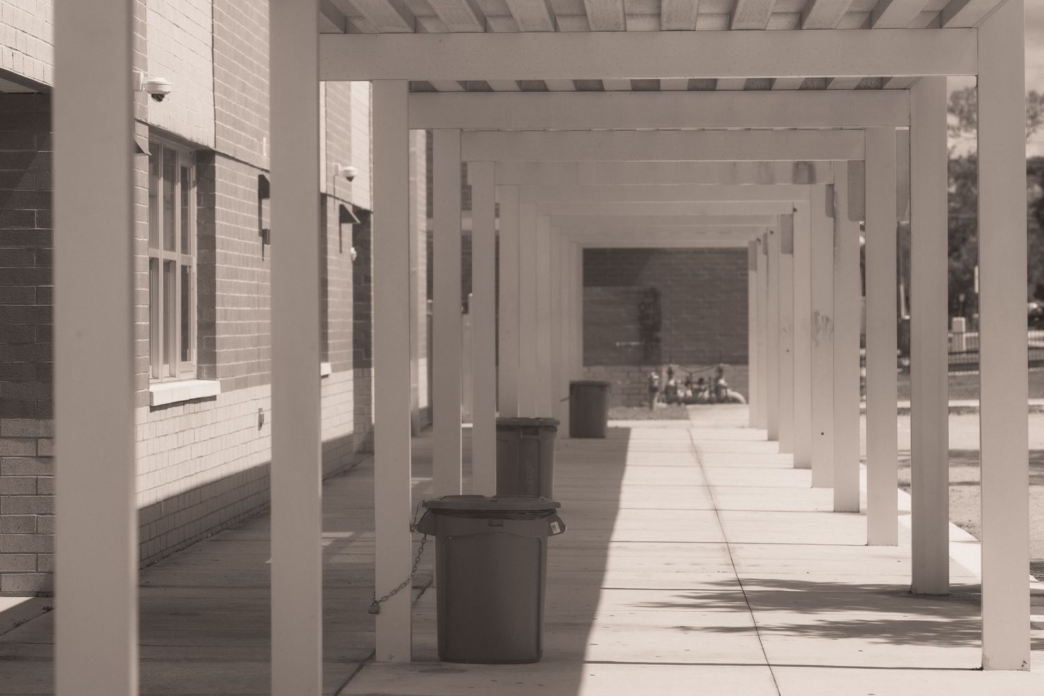 Sepia-toned covered walkway with white columns casting shadows on a concrete path beside a brick building.