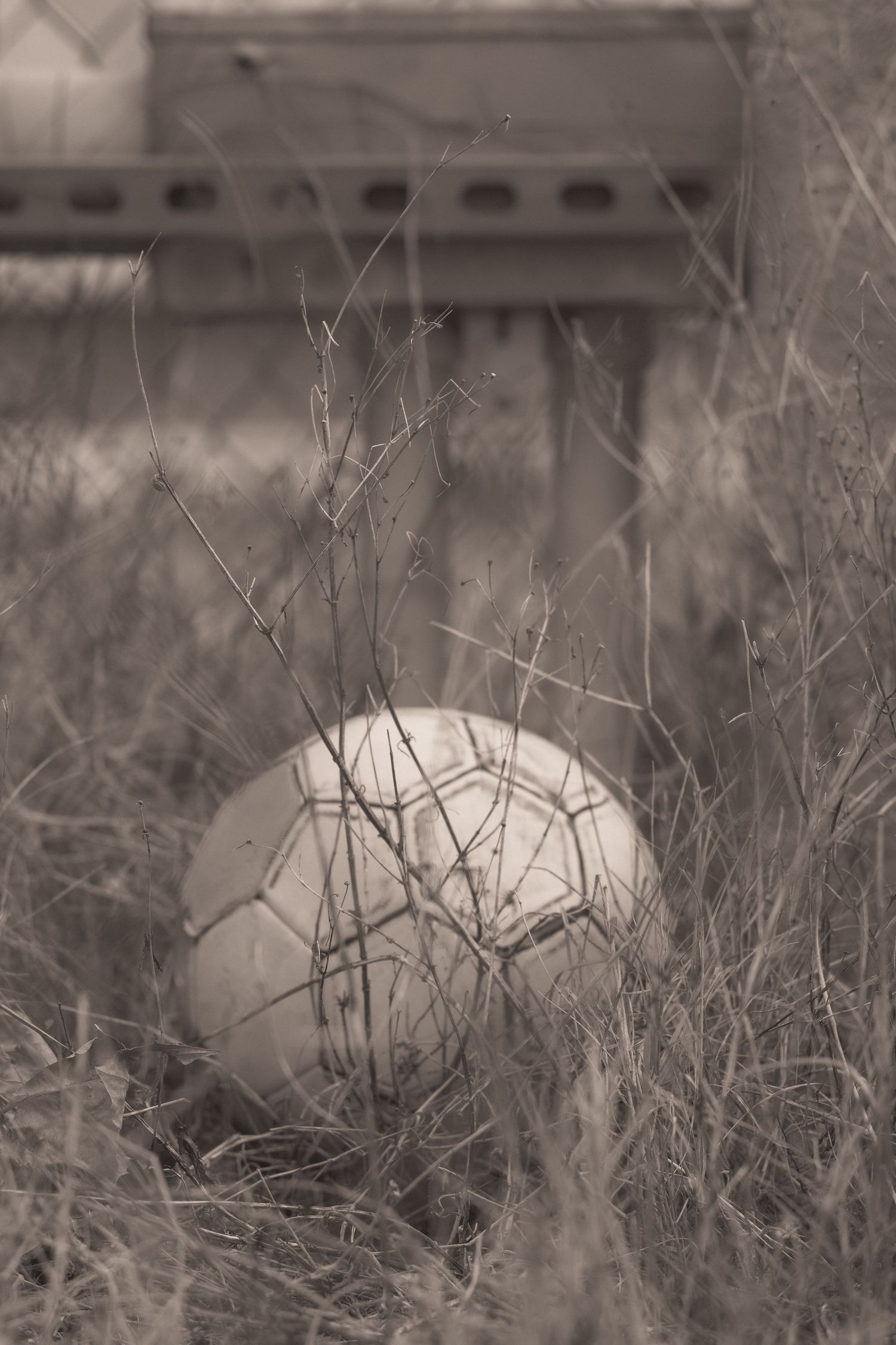 Vintage sepia photo of an old soccer ball resting in dry grass near a rusty metal structure.