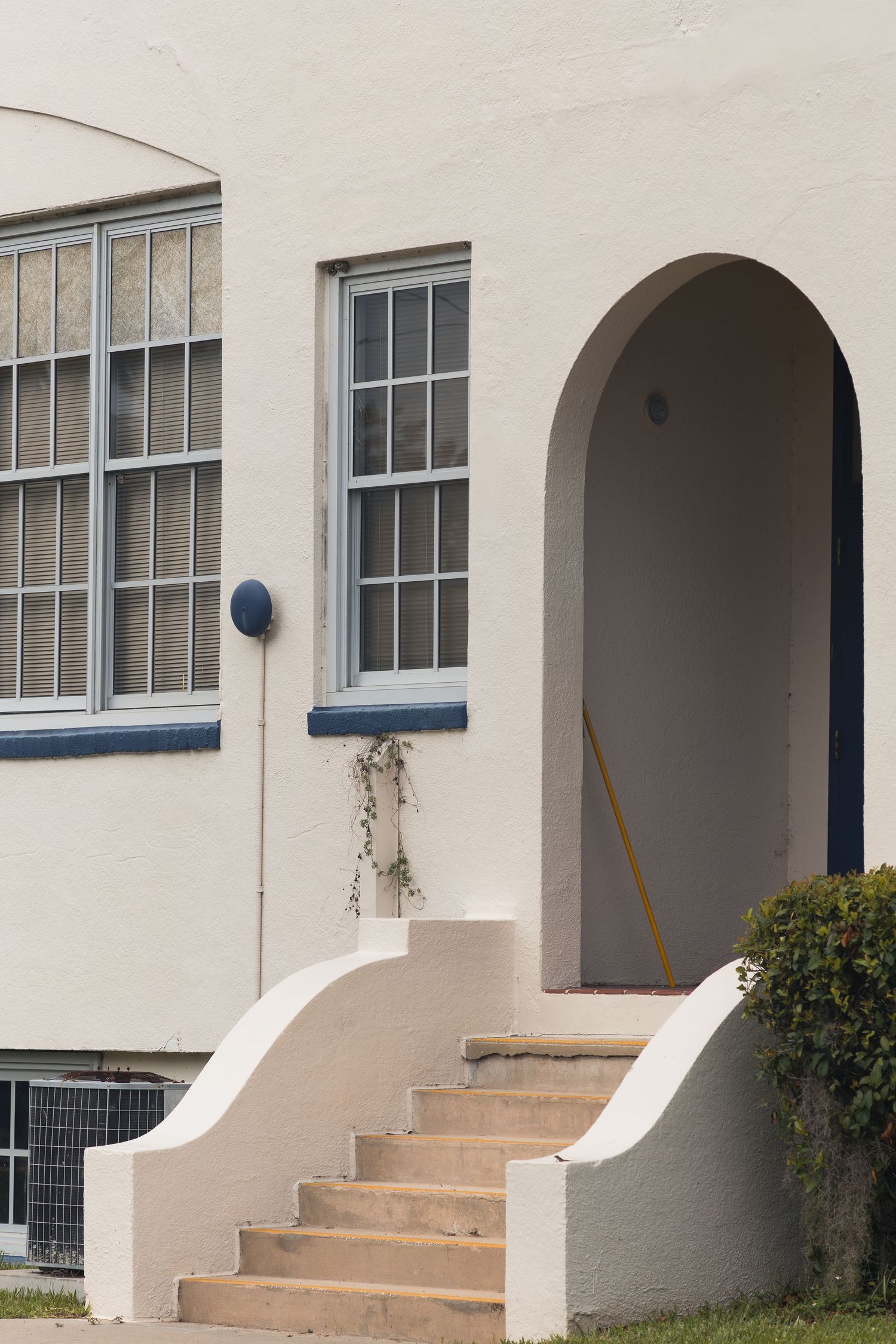 White Art Deco building entrance with arched doorway, steps, and blue-trimmed windows.