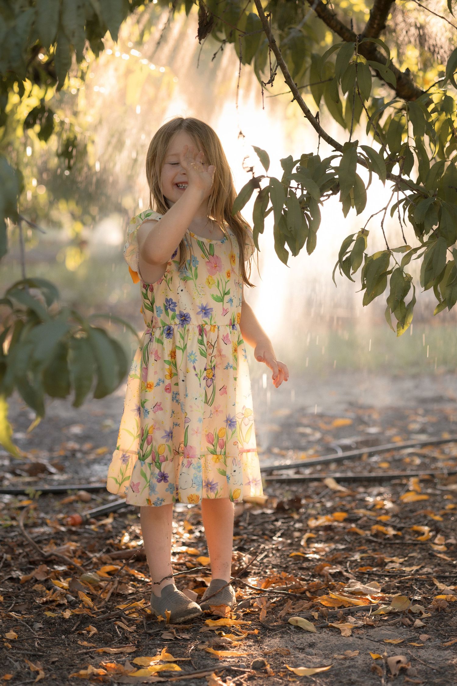 Young girl in floral dress playing under tree mist in golden sunlight, surrounded by autumn leaves.