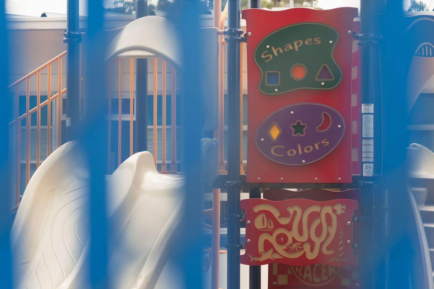 Colorful playground equipment with shapes and colors educational panel beside a white slide, viewed through blue fence rails.
