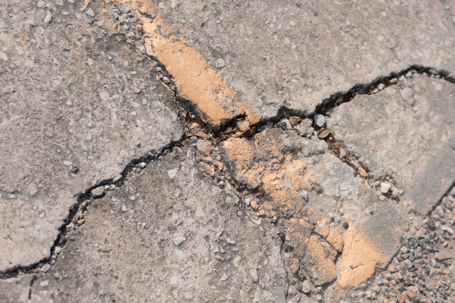 Close-up of cracked asphalt pavement with orange sand-filled fissures showing weathered road surface damage.
