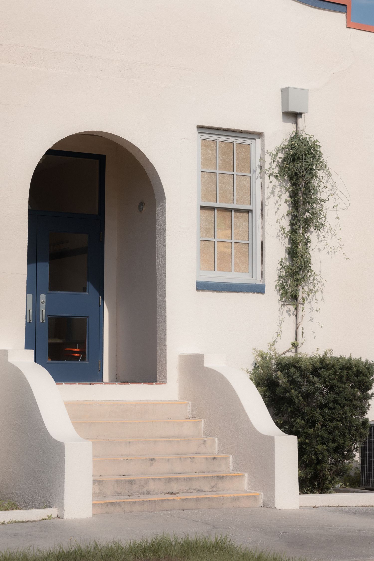 White stucco building entrance with arched blue door, stone steps, window with blue trim, and climbing vine plant.