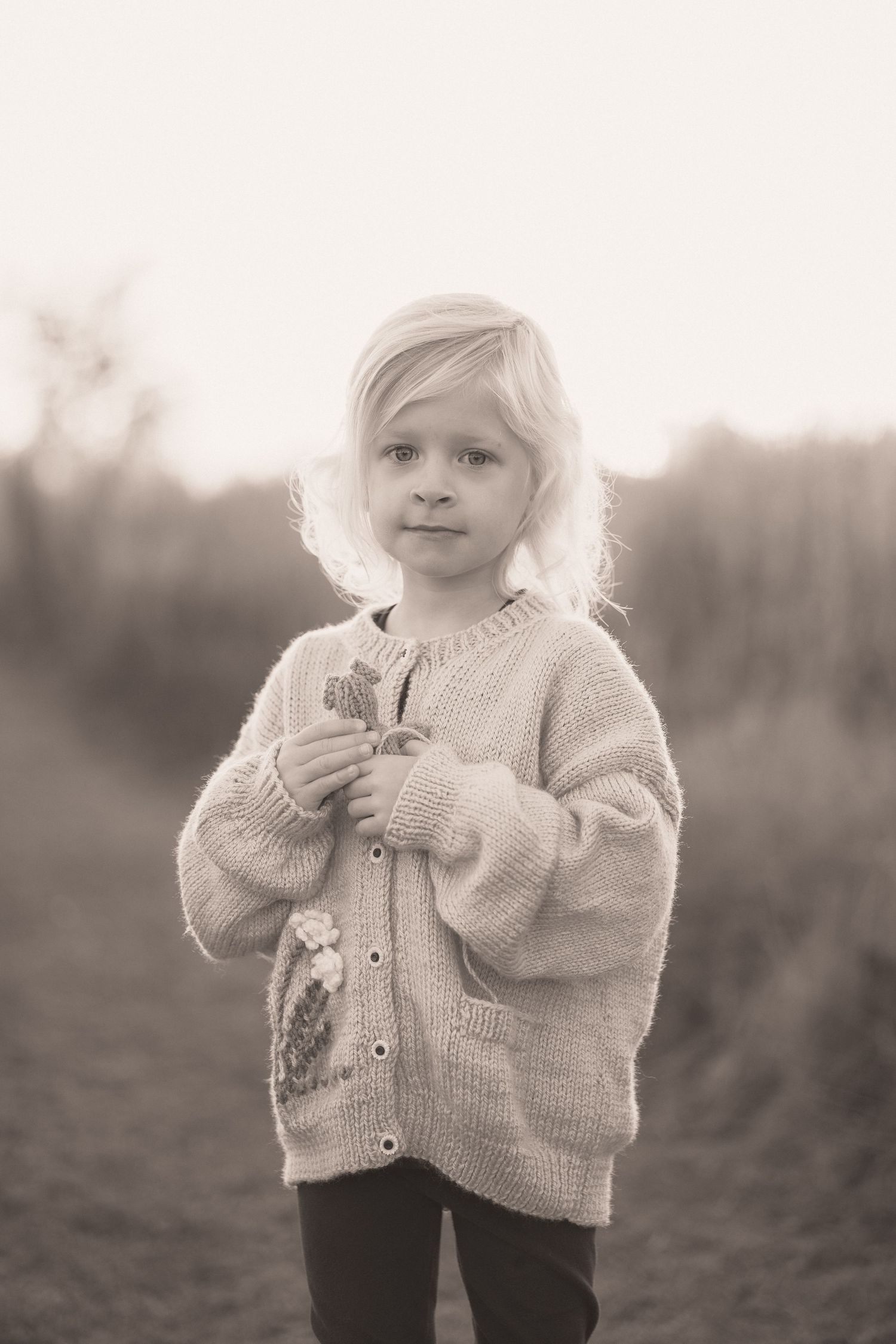 A young child in a cozy white knit cardigan stands outdoors in a sepia-toned photograph.