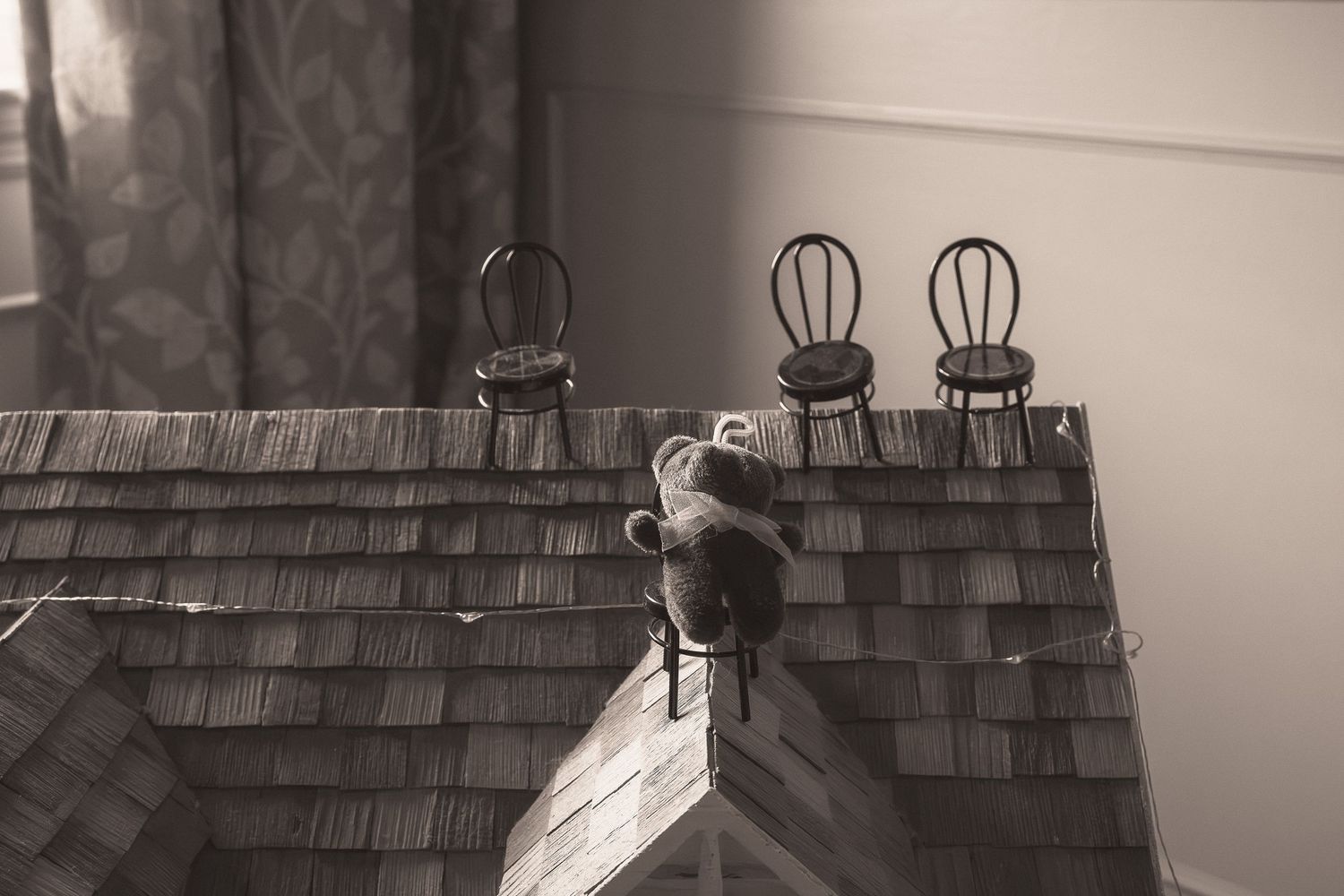 Black and white photo of a teddy bear sitting on a miniature shingled rooftop with small chairs in the background.