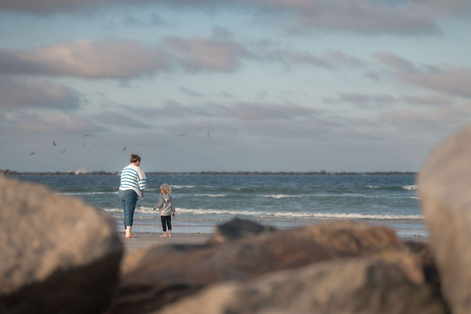 Two people walk along a rocky beach at sunset with waves crashing on the shore and cloudy skies overhead.