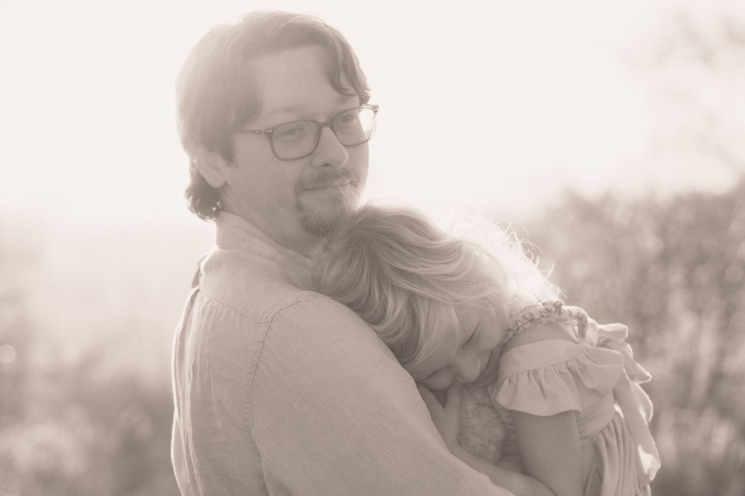 A man with glasses tenderly hugs a young girl outdoors in a warm sepia-toned portrait photo.