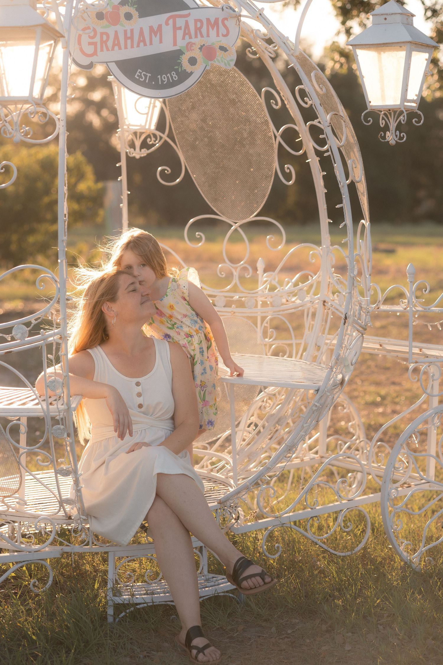 Mother and daughter share a tender moment on a decorative white iron carriage bench at Graham Farms during golden hour.