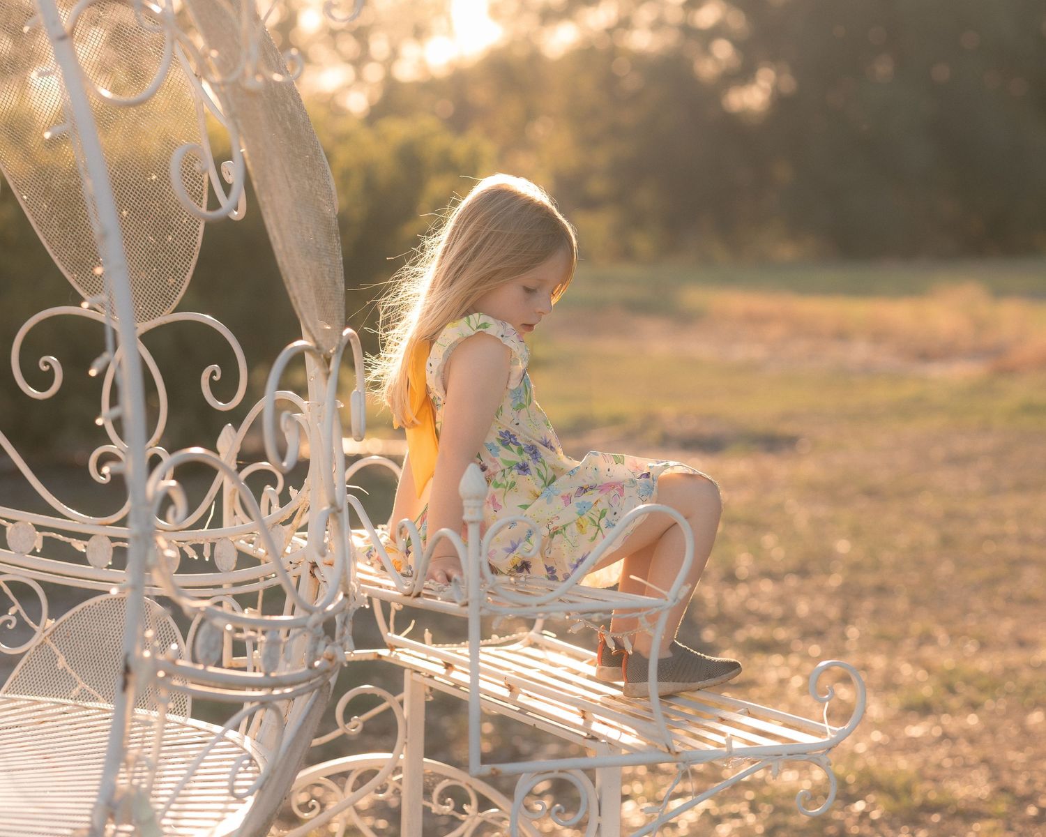 Young girl in floral dress sitting on ornate white metal bench in golden sunset light outdoors.