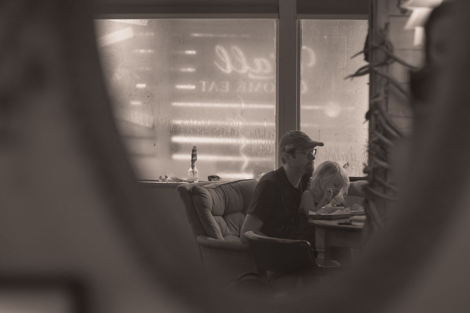Black and white photo of a man sitting alone in a dimly lit diner, reading near a glowing window at night.