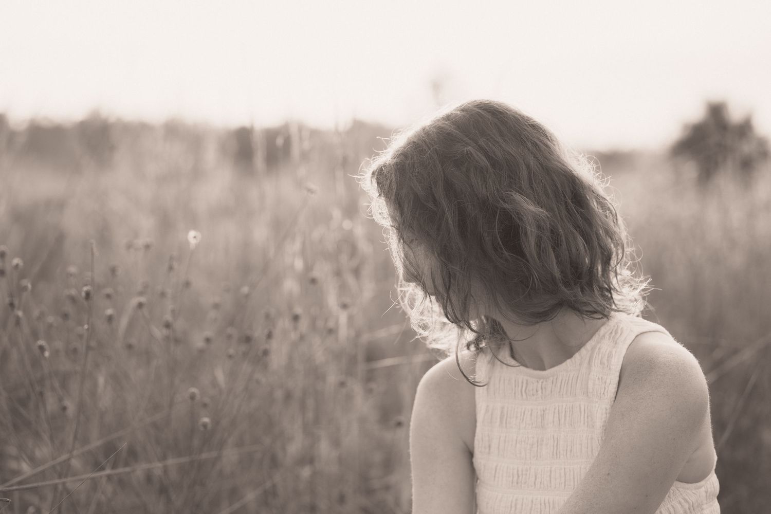 Black and white photo of a young girl in a white dress standing in a sunlit meadow, looking down thoughtfully.