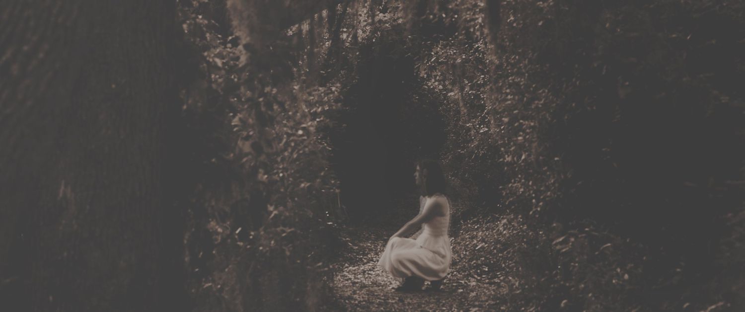 A solitary figure in white sits at the entrance of a dark, moss-covered cave surrounded by autumn foliage.
