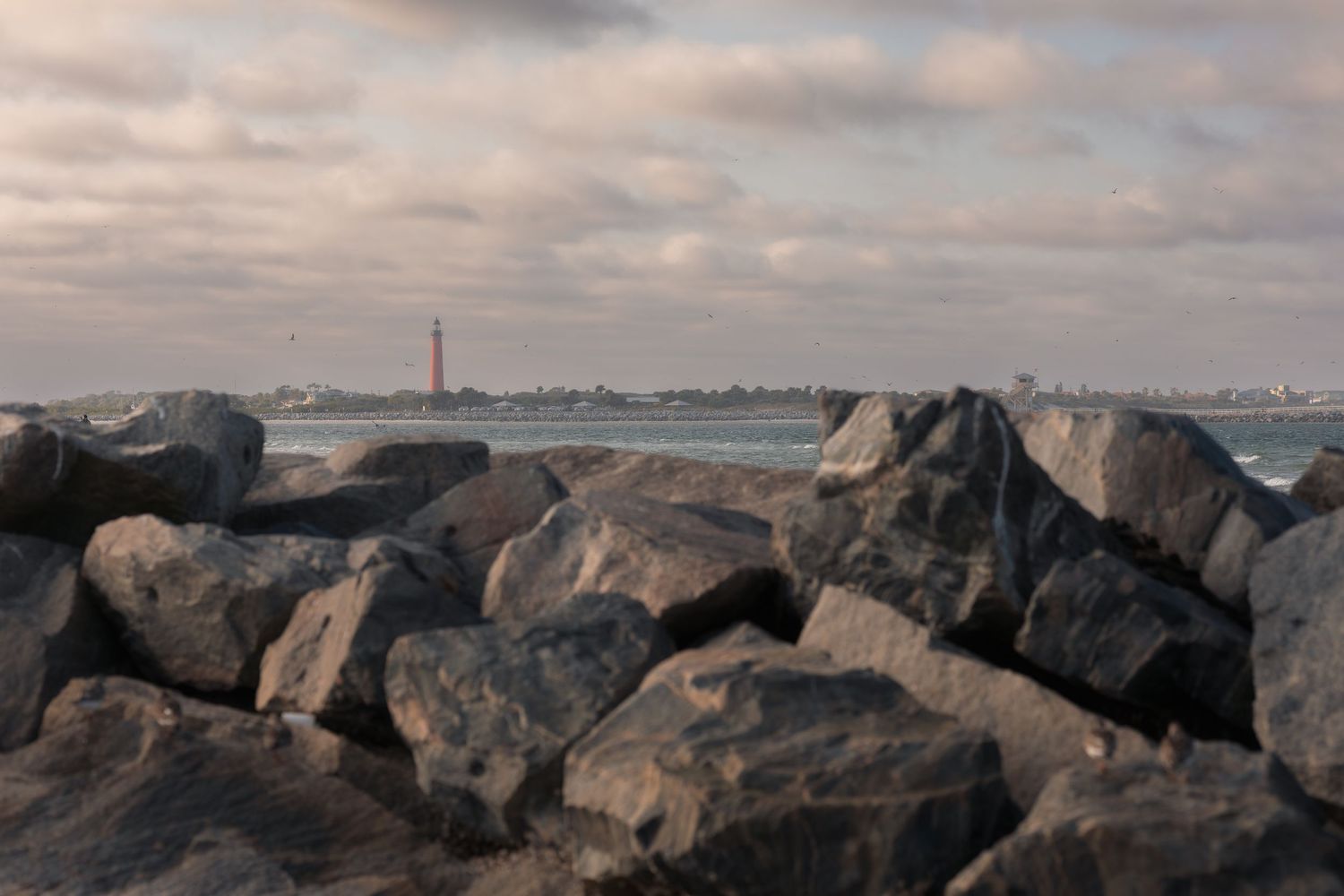 Large rocks line a rocky shoreline at sunset with calm waters and distant land visible on the horizon.