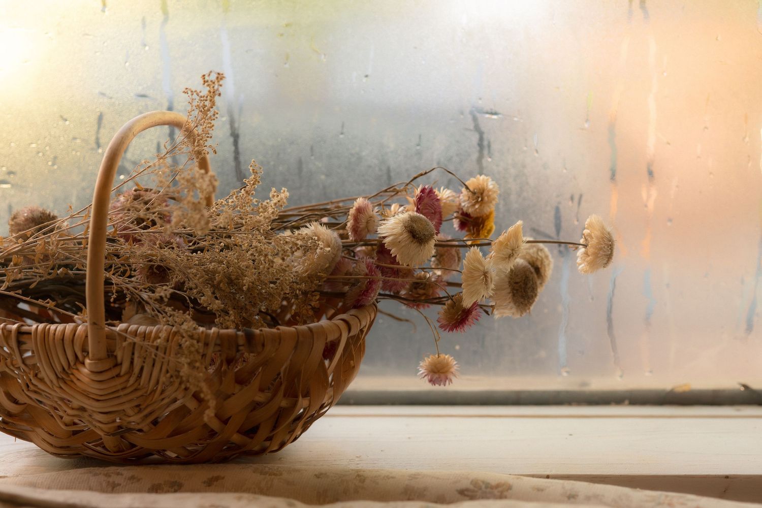 A wicker basket filled with dried wildflowers and herbs sits on a windowsill beside a foggy, moisture-covered window.